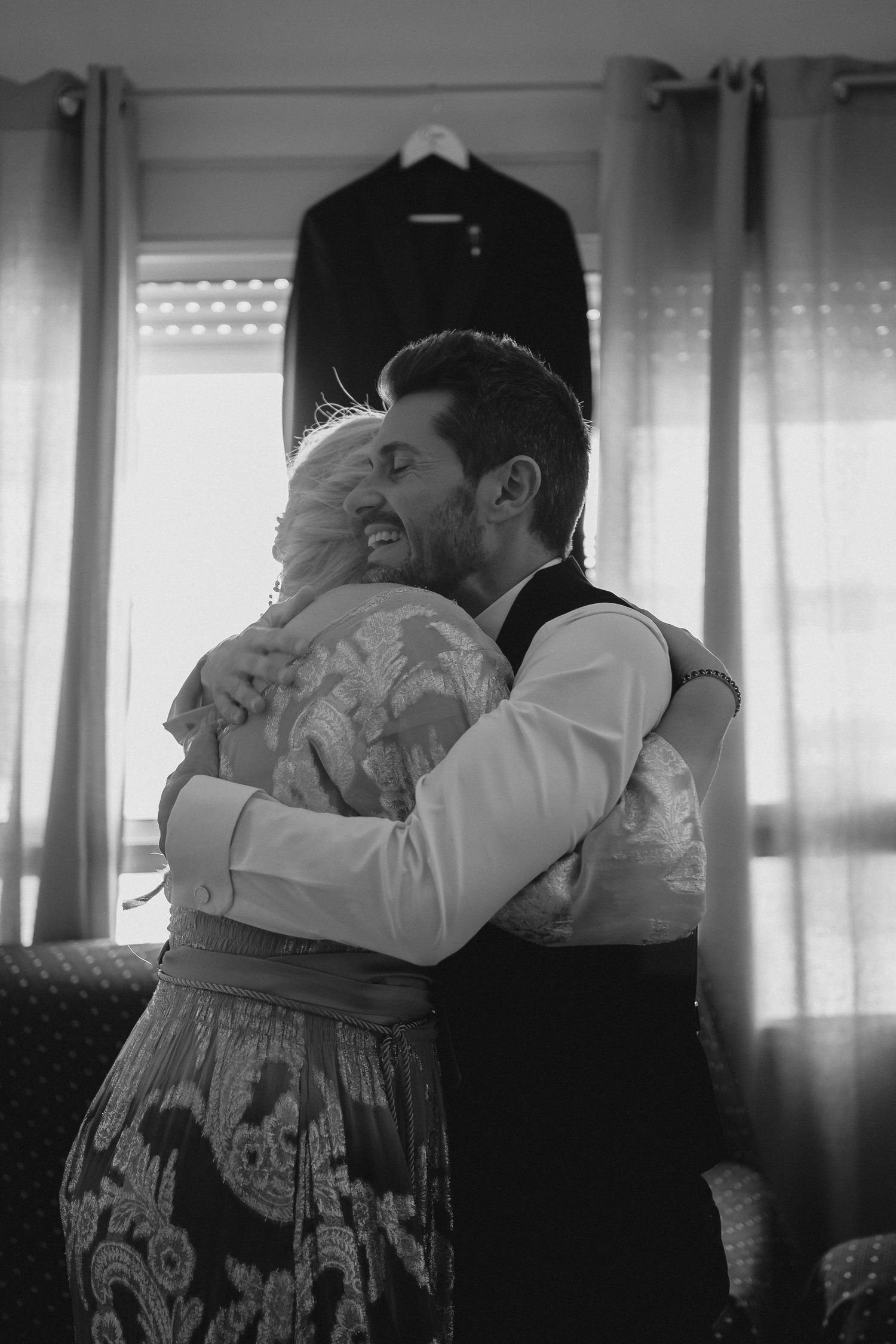 A black and white photo of a groom in a vest embracing an older woman in a patterned dress.