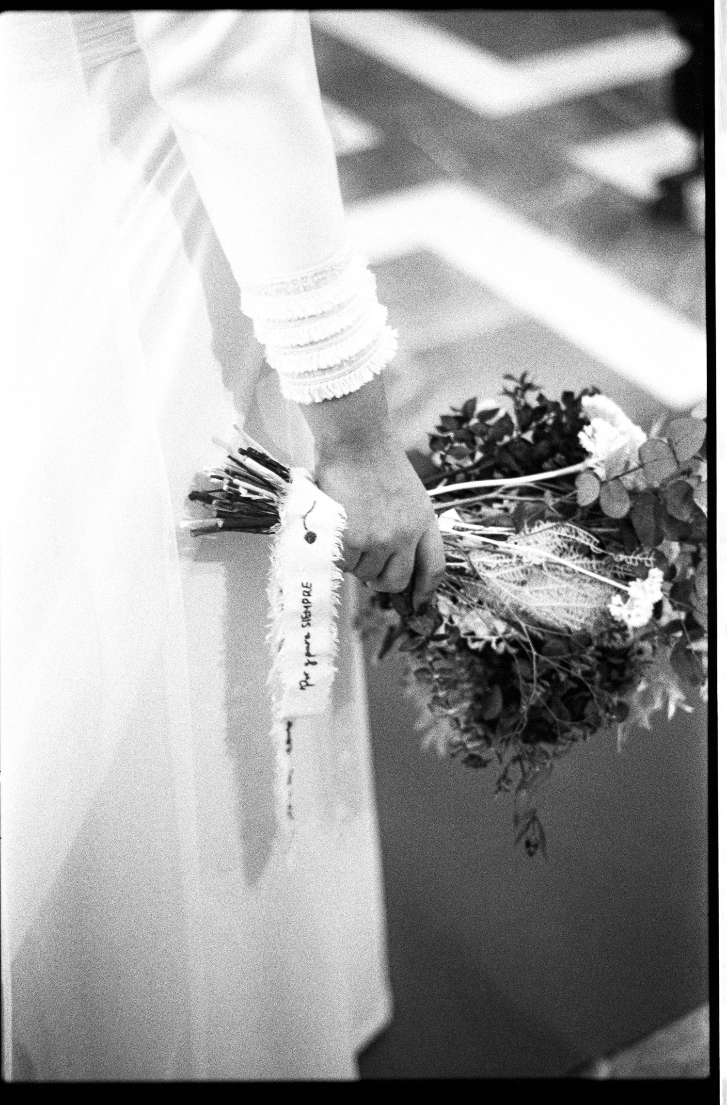Black and white close-up of bride holding small wildflower bouquet with ribbon tag at wedding ceremony.