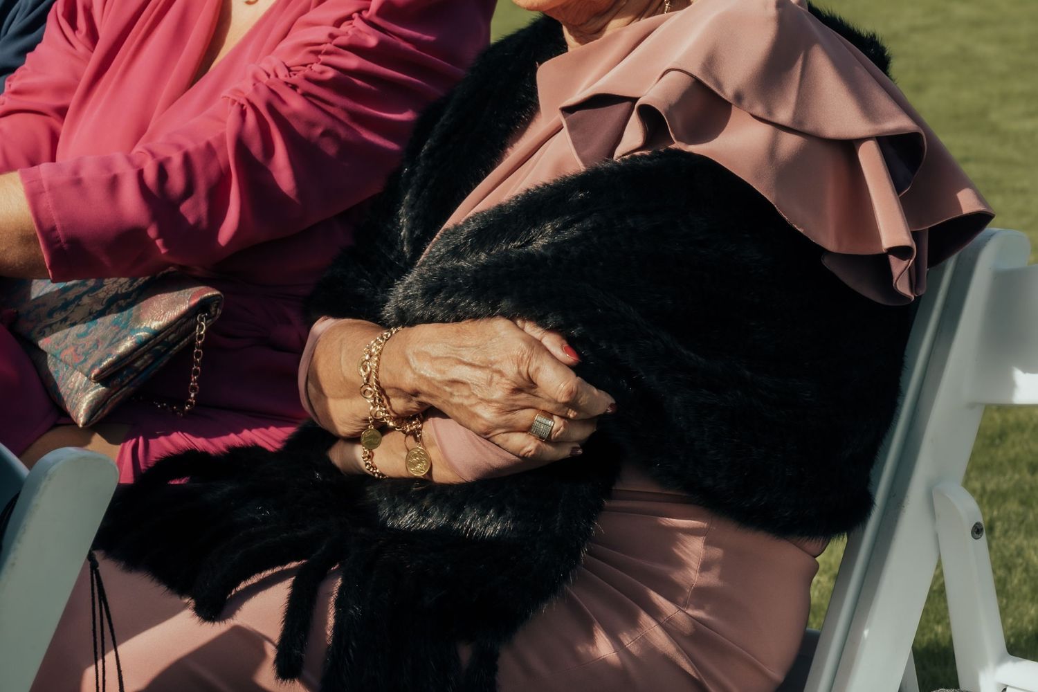 Two elderly women in pink and magenta outfits holding hands with gold bracelets at an outdoor wedding ceremony.