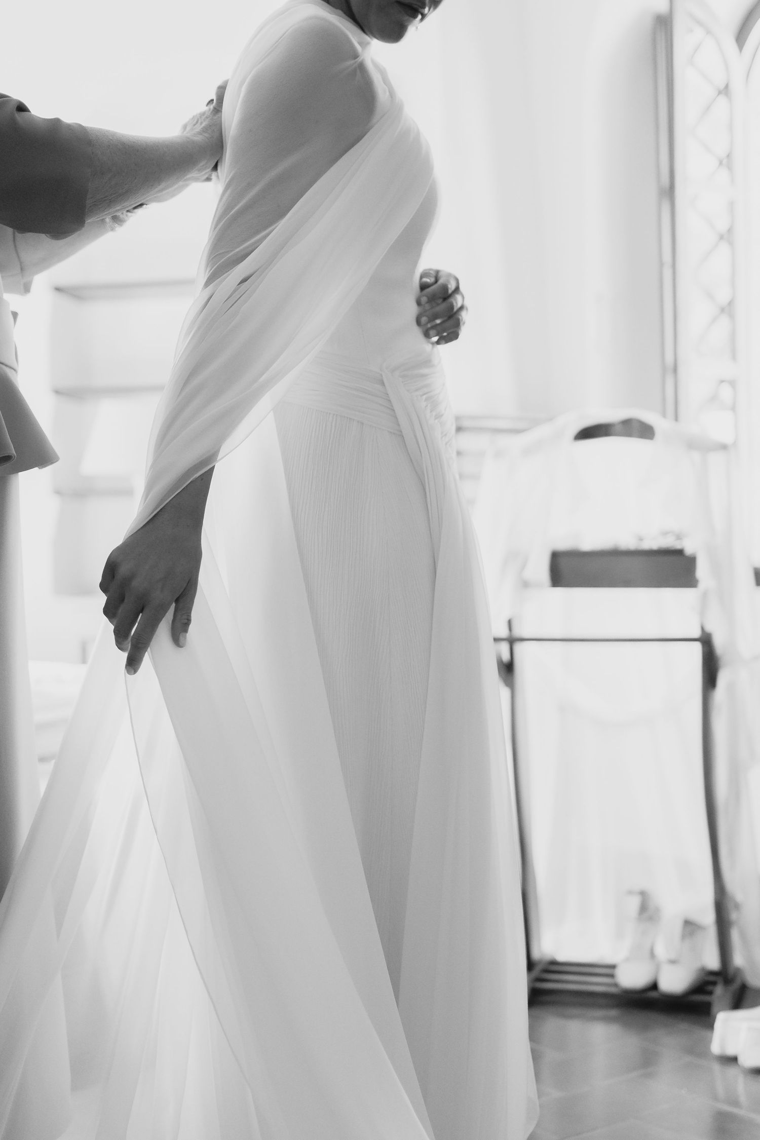 Black and white image of assistant fastening bride into elegant white wedding dress.