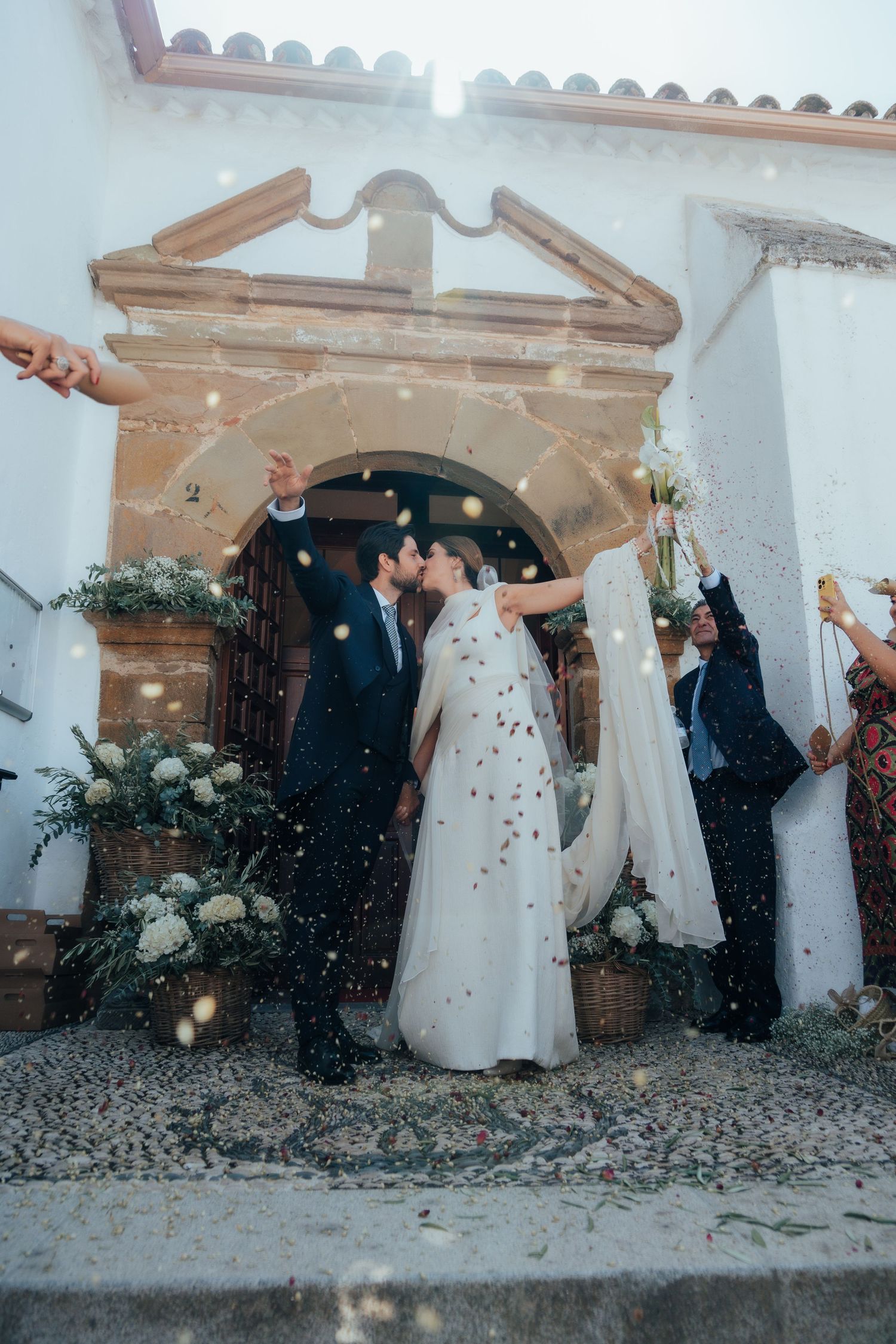 Newlyweds sharing a kiss outside a white Spanish church as guests throw confetti in celebration.