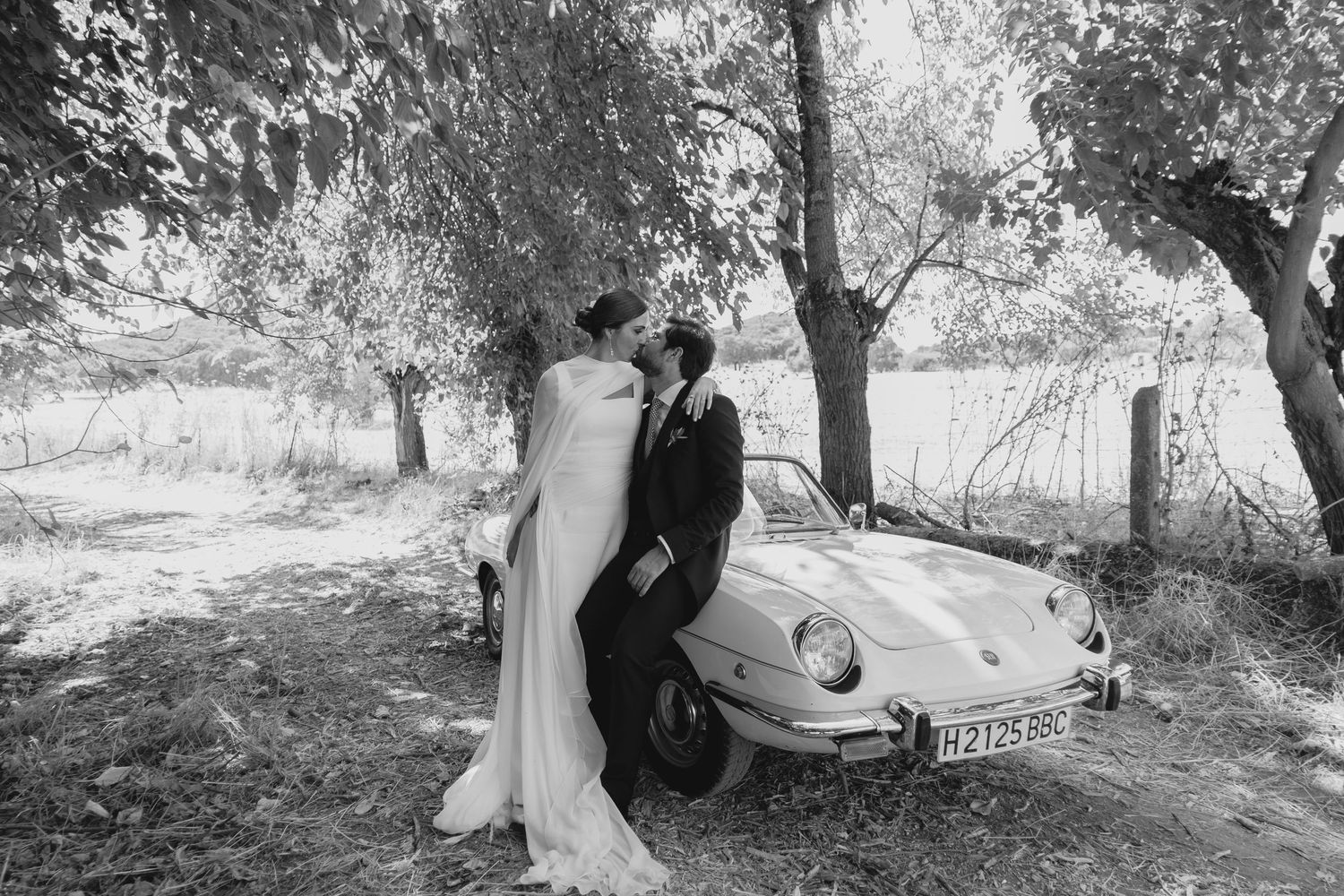 Bride and groom share a romantic moment beside a vintage car under trees in a black and white wedding photo.