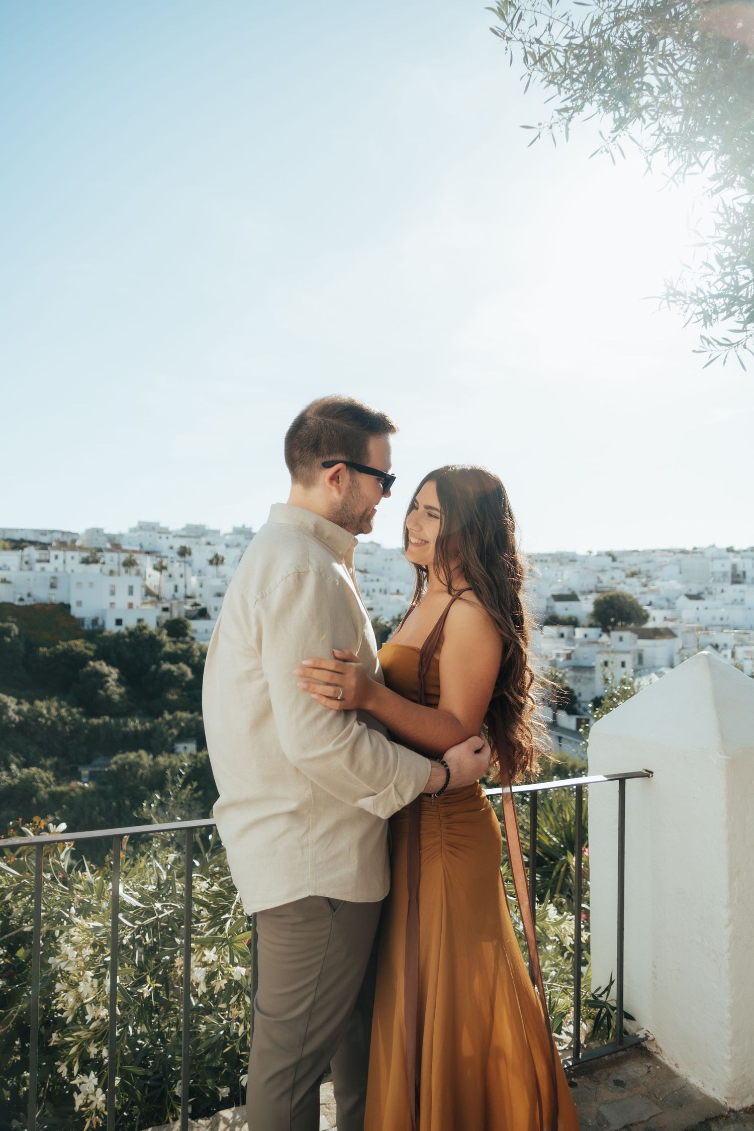 Romantic couple embracing on a sunlit balcony with white-washed Mediterranean village in the background.