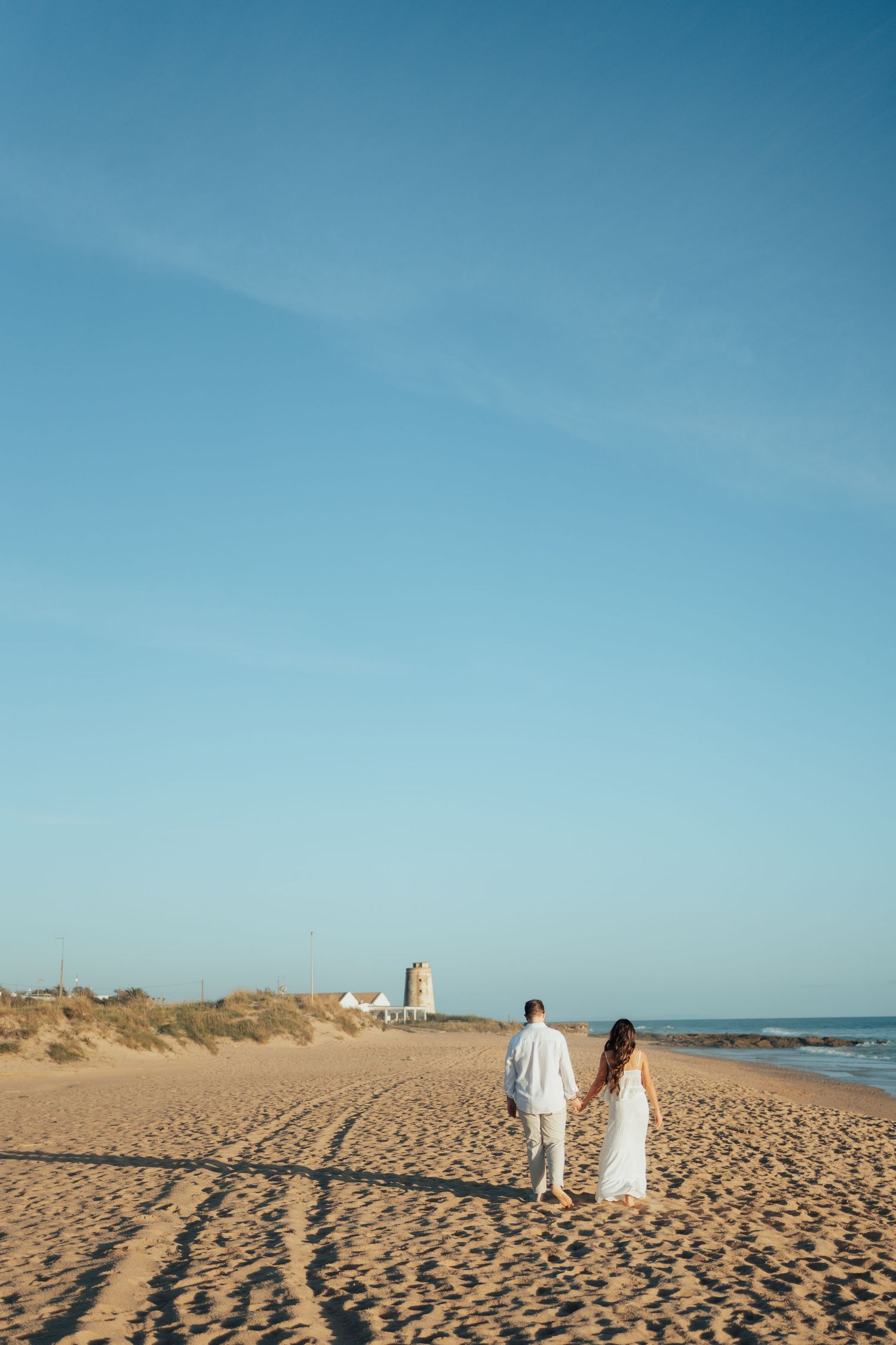 Couple in white outfits holding hands while walking on a sandy beach with blue sky and ocean in the background.
