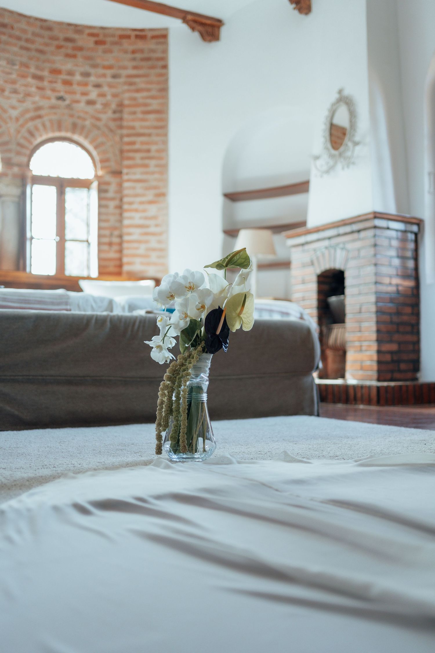 White orchid and anthurium bouquet in glass vase in rustic brick-walled bedroom interior.