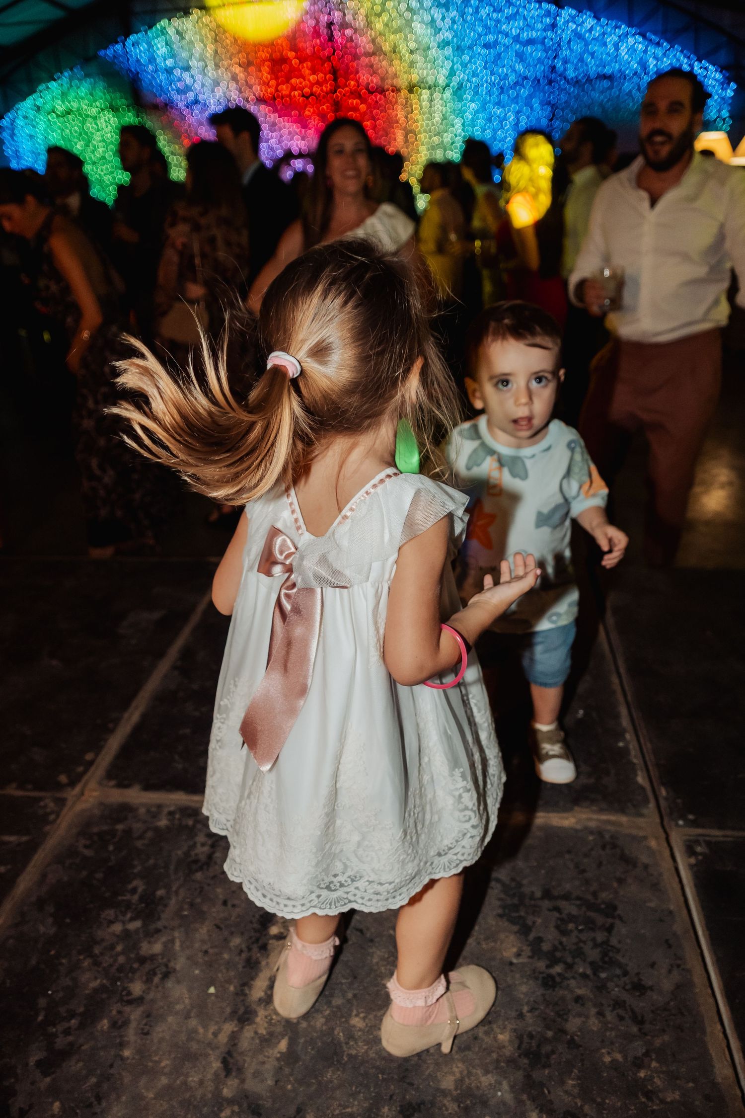 Young girl in white dress dancing energetically with toddler under colorful lights at wedding reception.