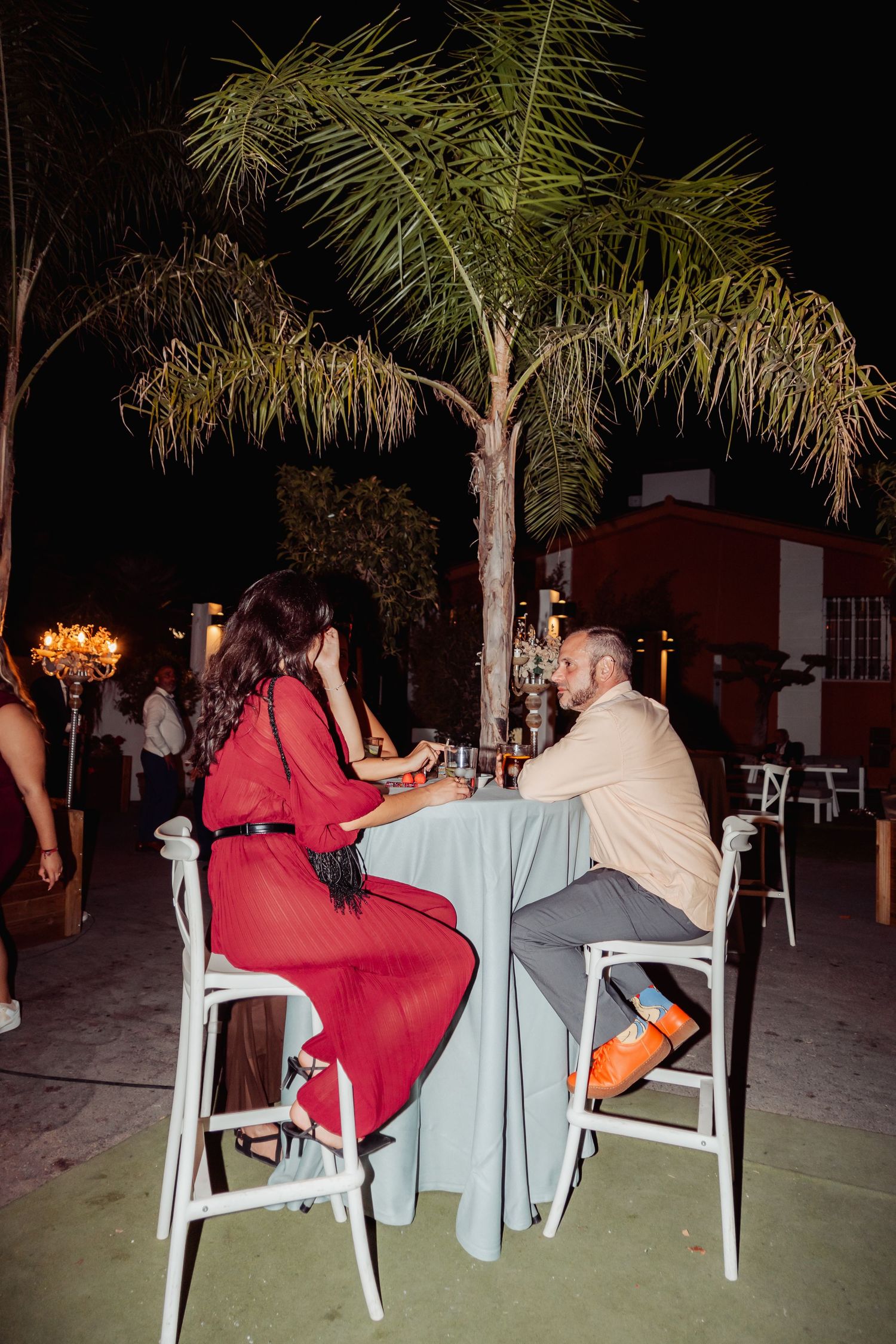 Couple in elegant attire sharing drinks at a candlelit outdoor table during a night wedding reception.