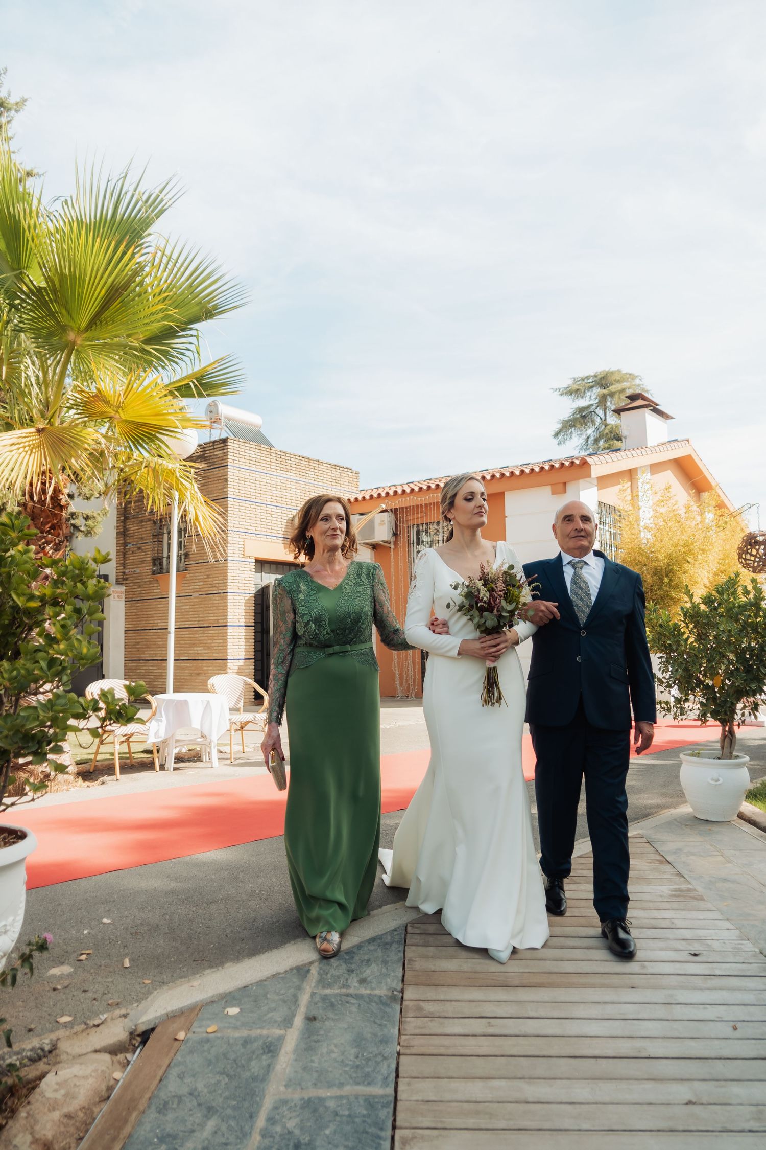 Bride walks down a red carpet path escorted by her parents on a sunny wedding day.