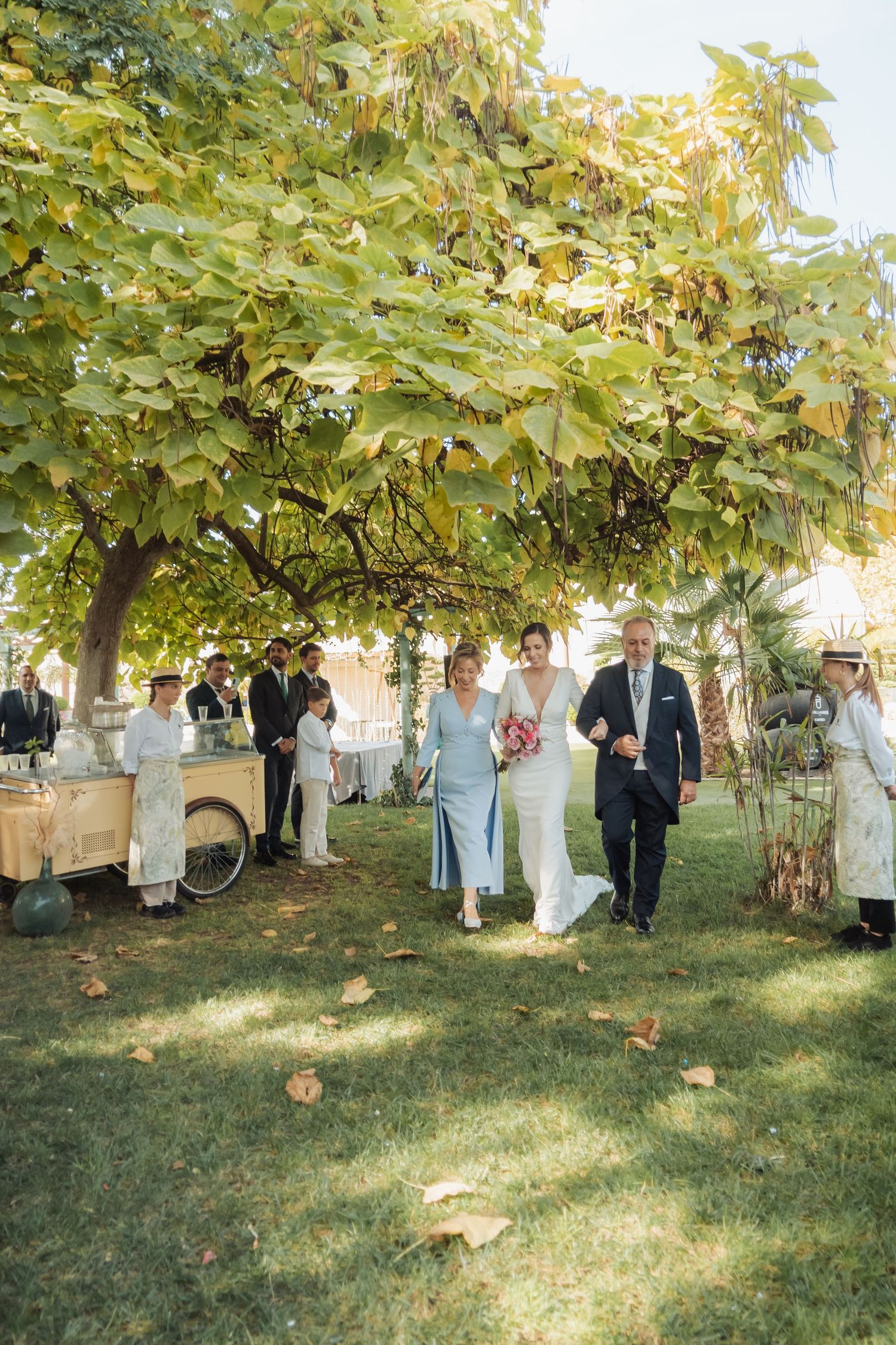Bride in white gown walks with parents under large tree at outdoor wedding.