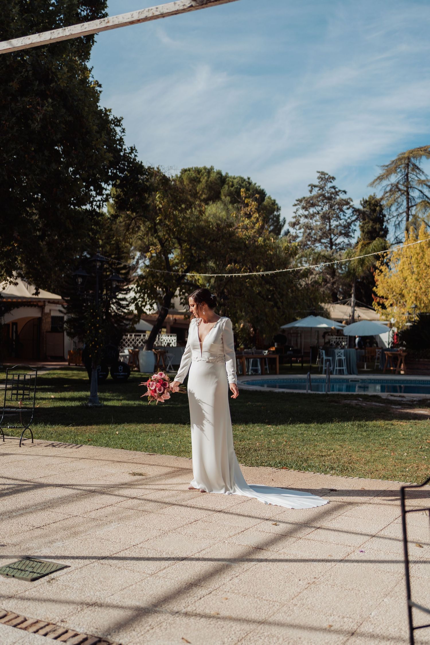 Bride in sleek white long-sleeve wedding gown with train standing in sunny garden venue.