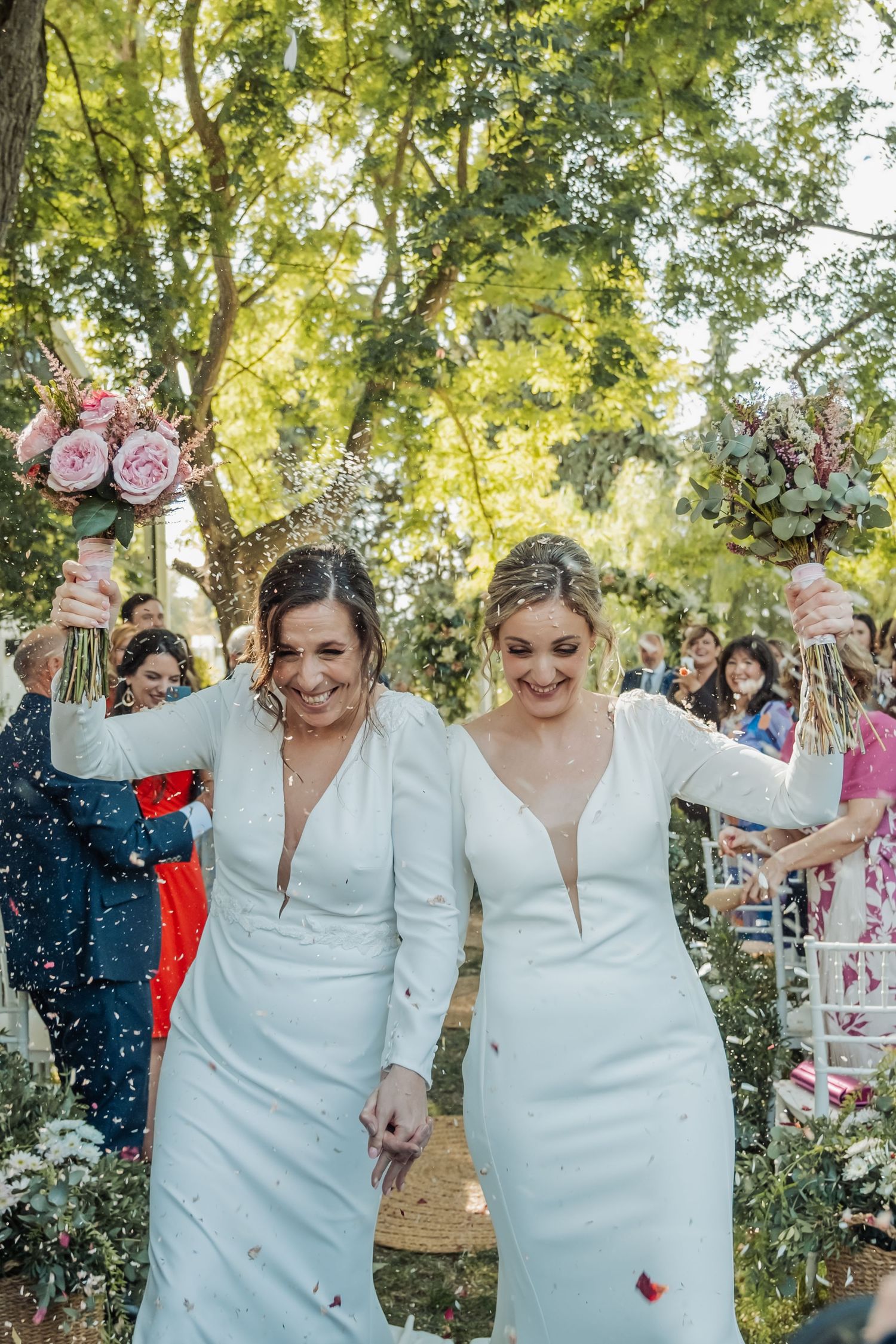 Two brides in white dresses holding colorful bouquets walking through confetti shower at outdoor wedding.