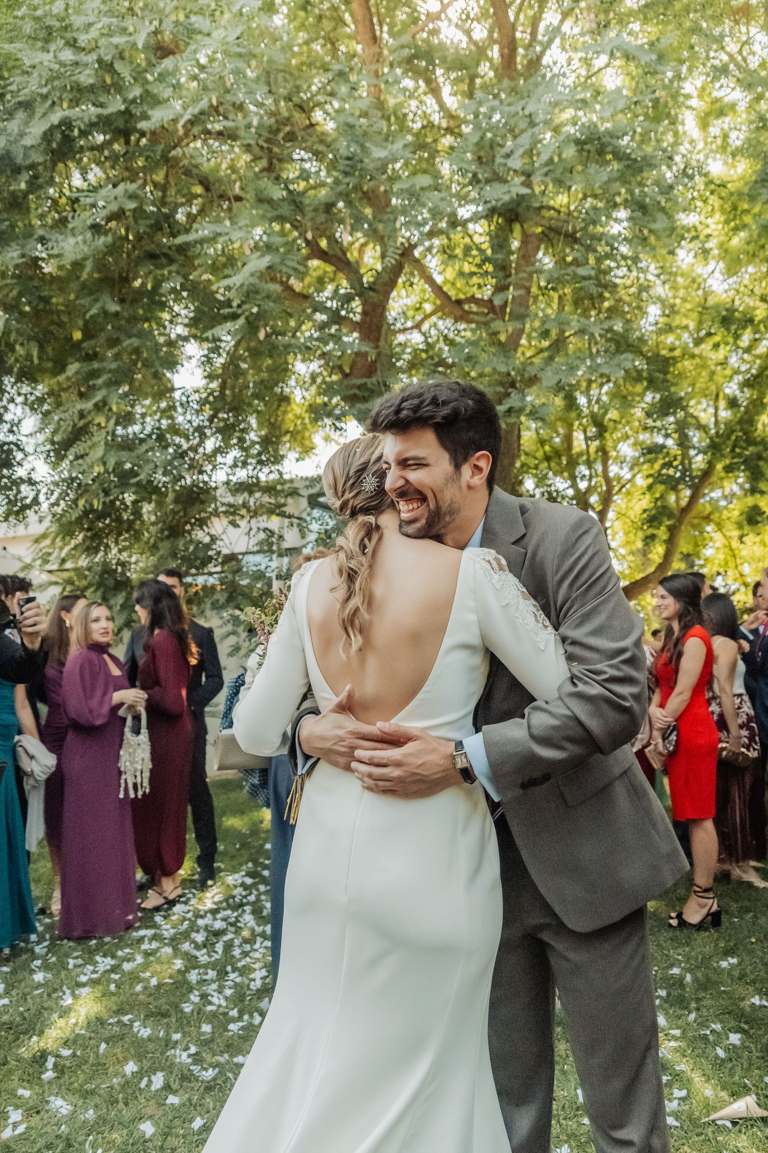 Groom in gray suit embraces smiling bride in backless white dress at outdoor wedding celebration.