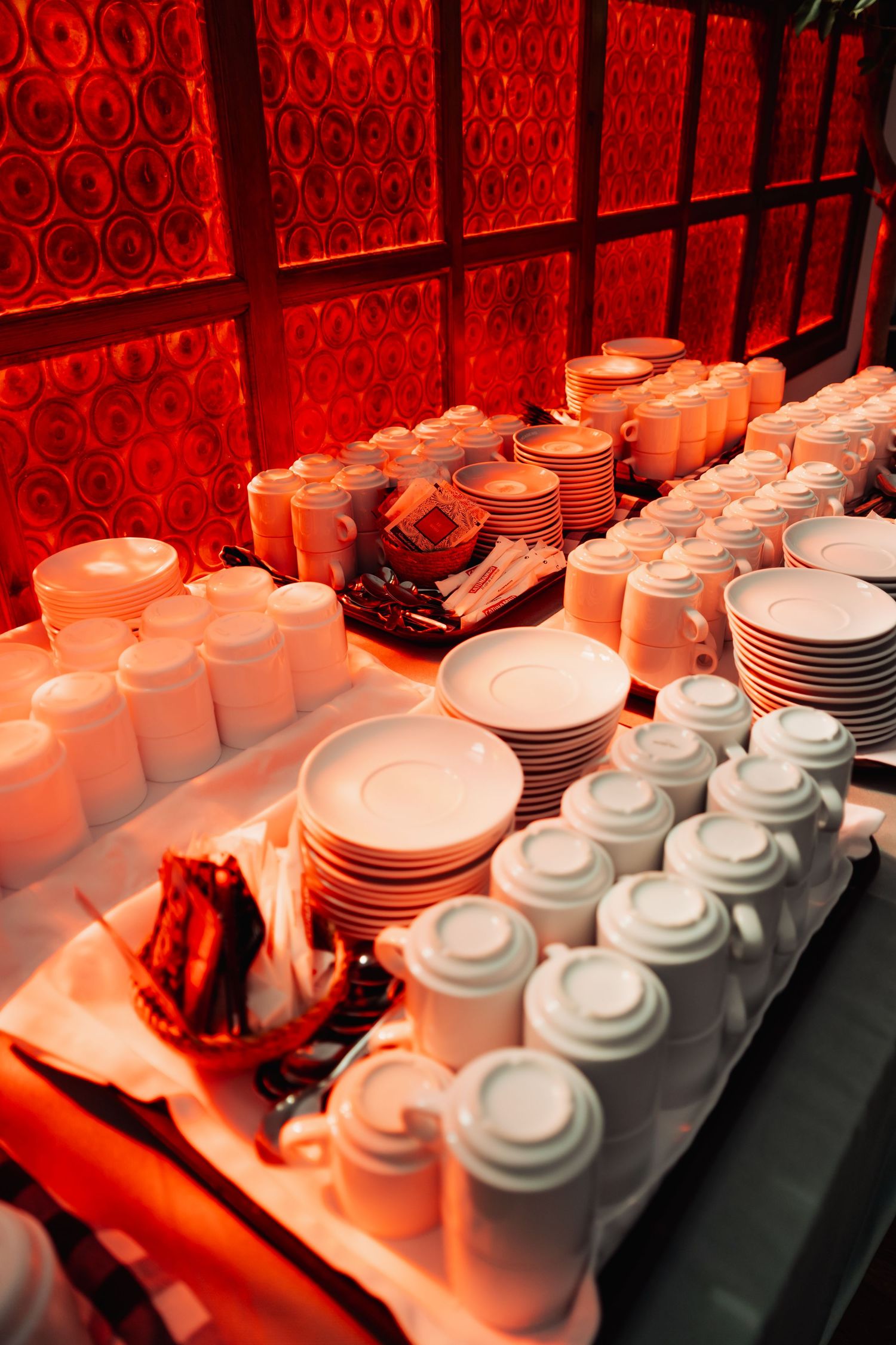 White ceramic dishes, cups, and teapots arranged on a buffet table under warm red ambient lighting.