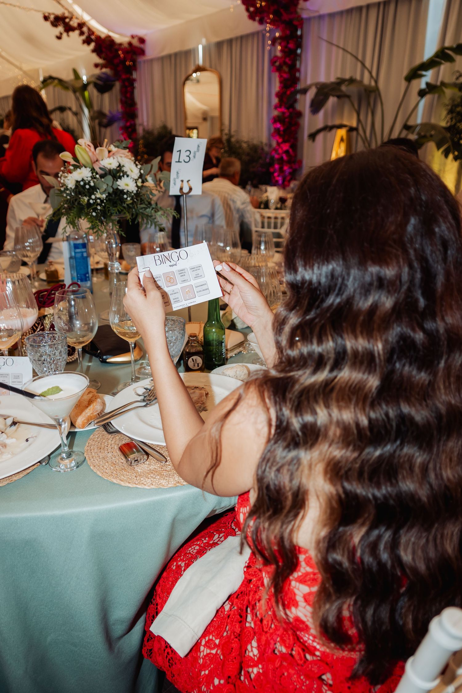Wedding guest in red dress reads a bingo card at table 13 during a festive reception dinner.