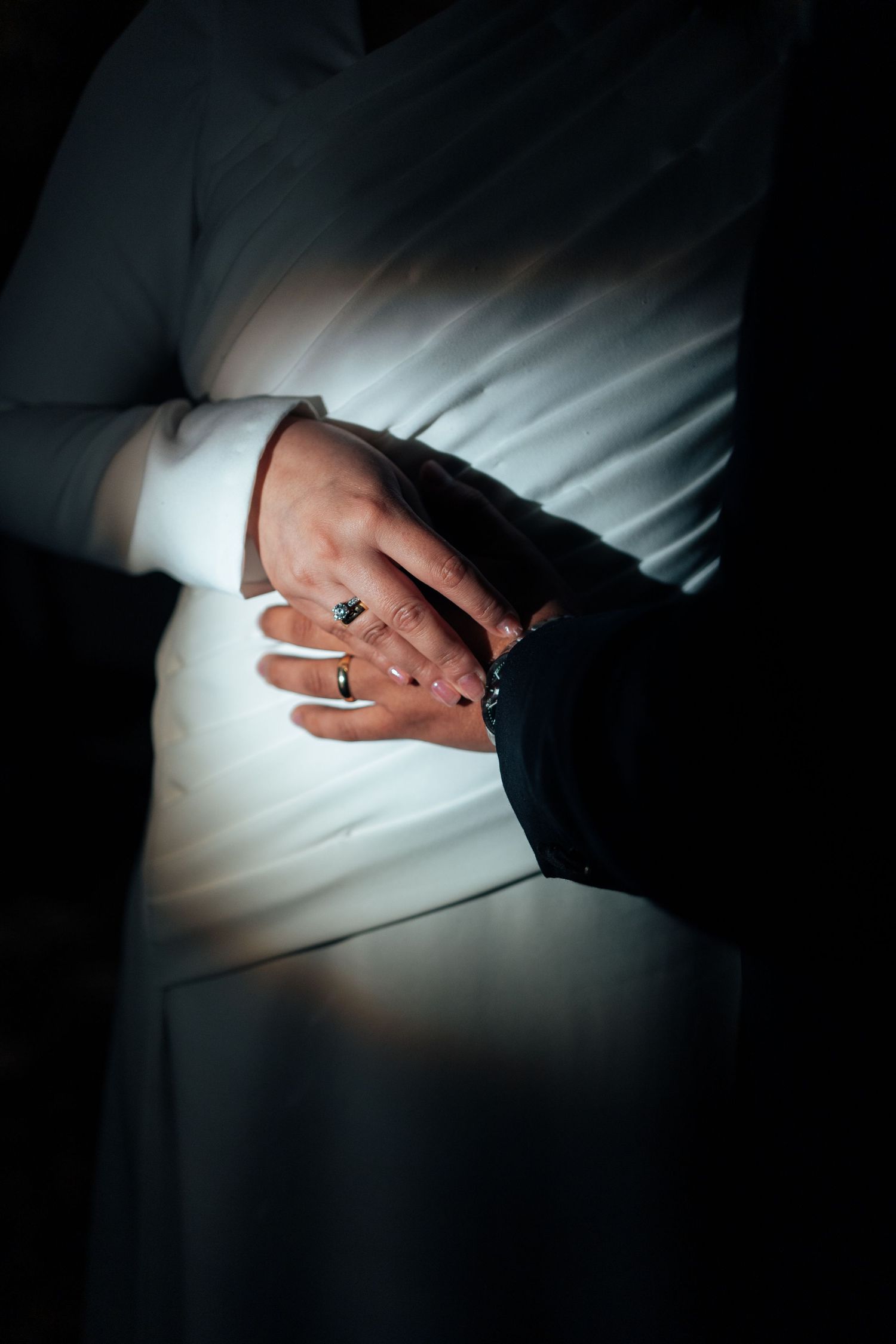 Close-up of bride and groom holding hands showing wedding rings in dramatic dark lighting.