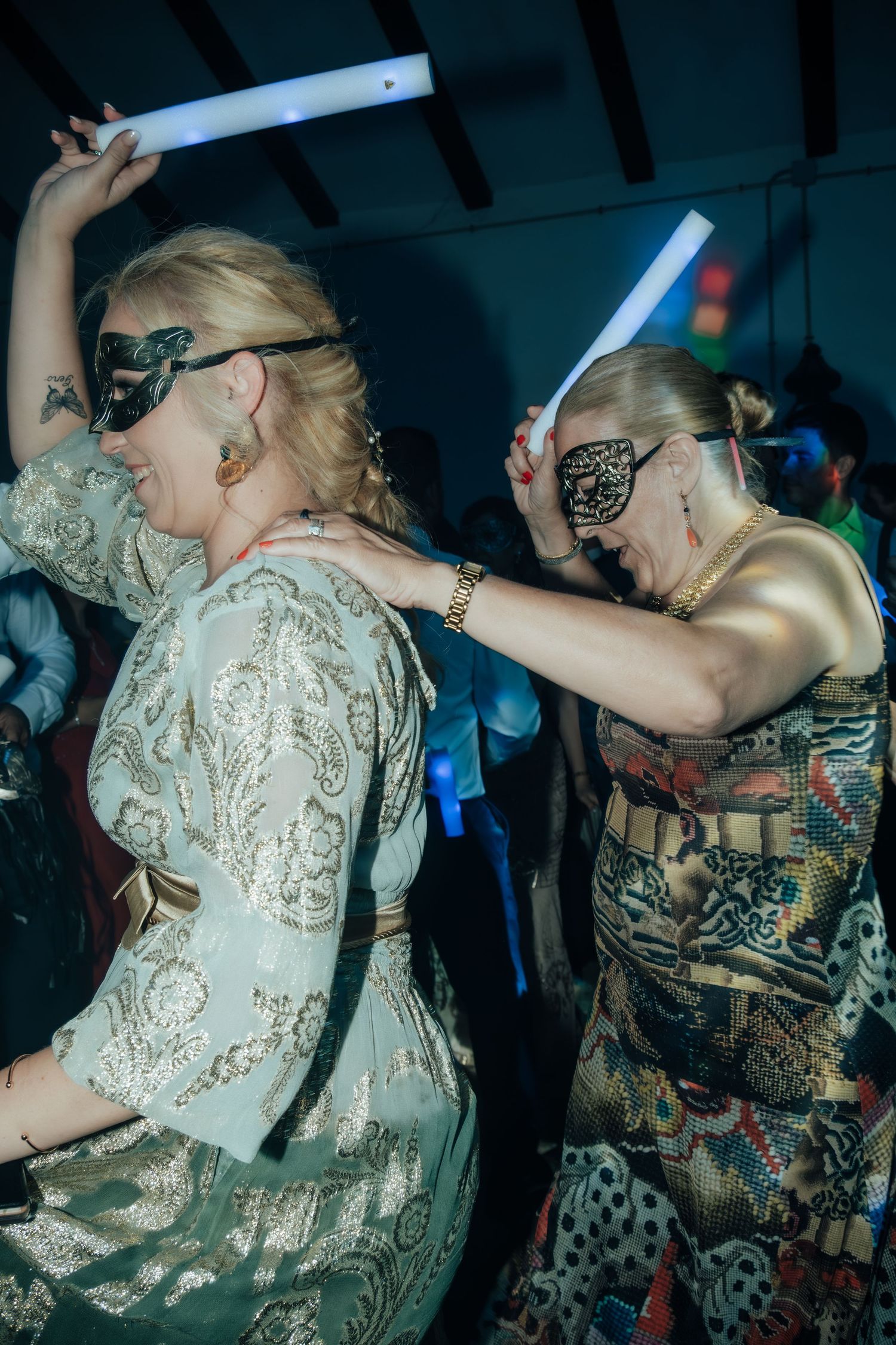 Two women in elegant dresses and masquerade masks dance together at a glamorous masquerade party with glowing neon lights.