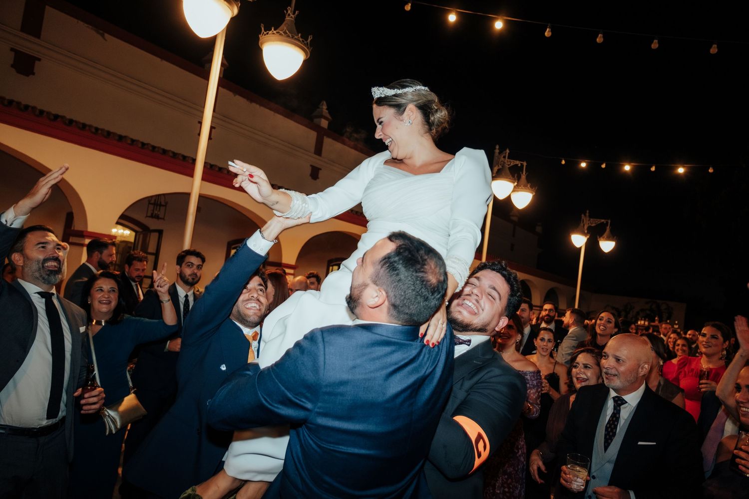 Bride lifted by cheering wedding guests during energetic outdoor evening reception celebration.