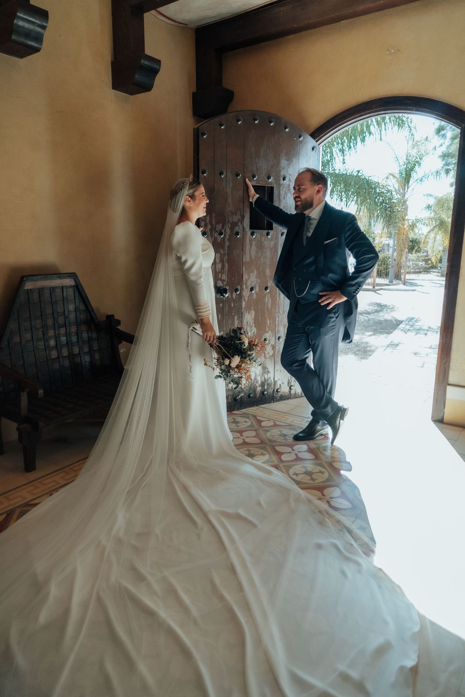 Bride in white gown with long veil holds bouquet as groom opens ornate wooden arched door.
