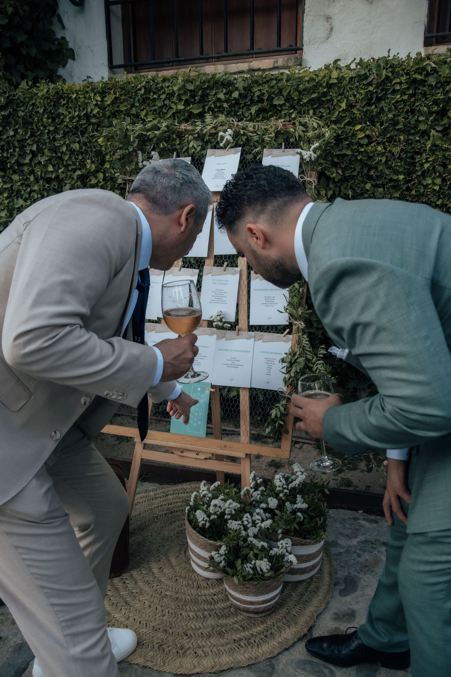 Two male wedding guests in suits leaning in to read seating chart on wooden easel.