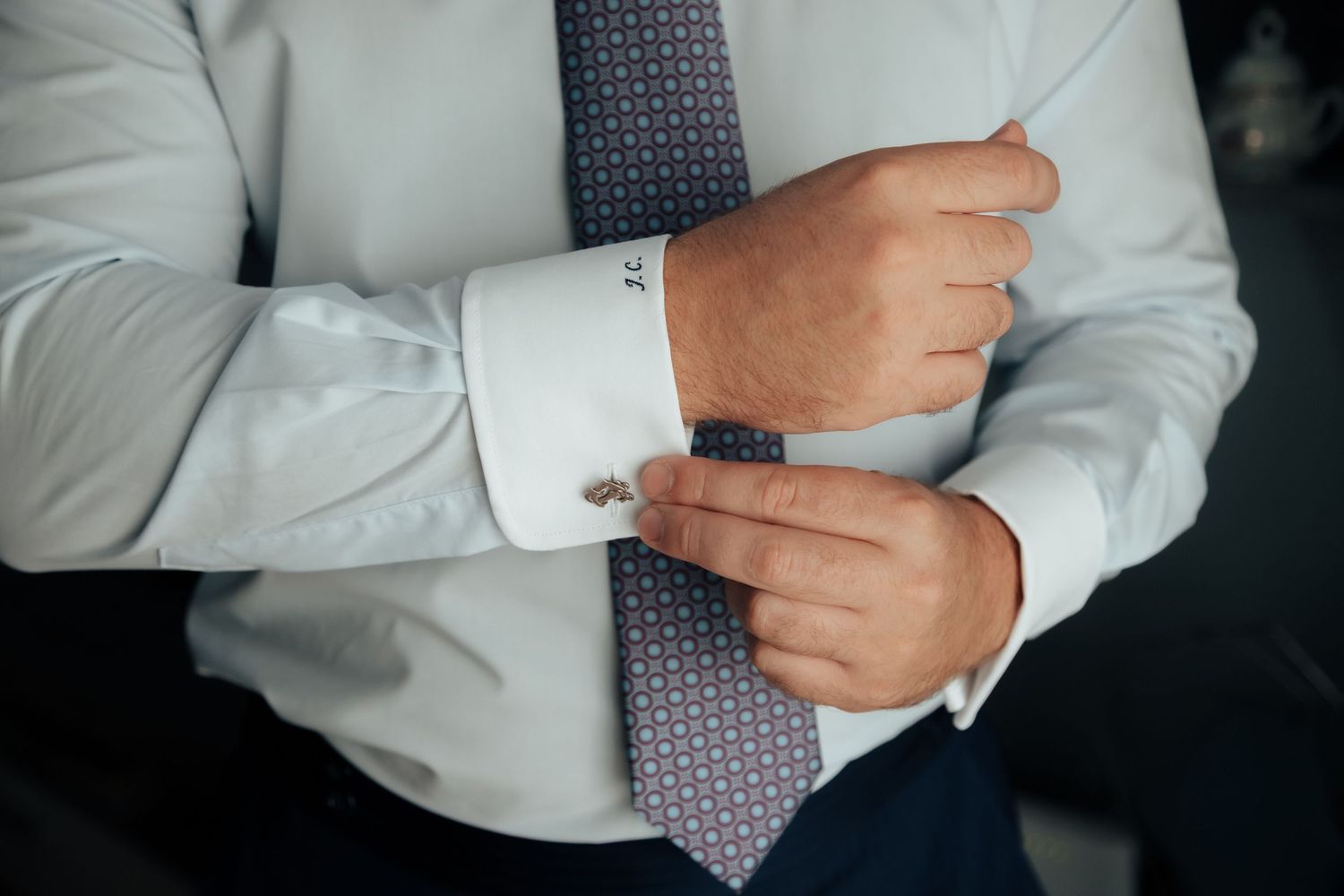 Groom in white dress shirt and patterned tie fastening silver cufflinks on his sleeve.
