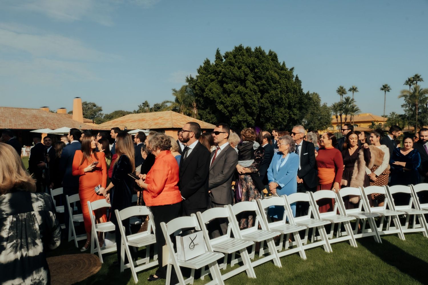 Wedding guests standing and watching outdoor ceremony seated in white folding chairs on green lawn.
