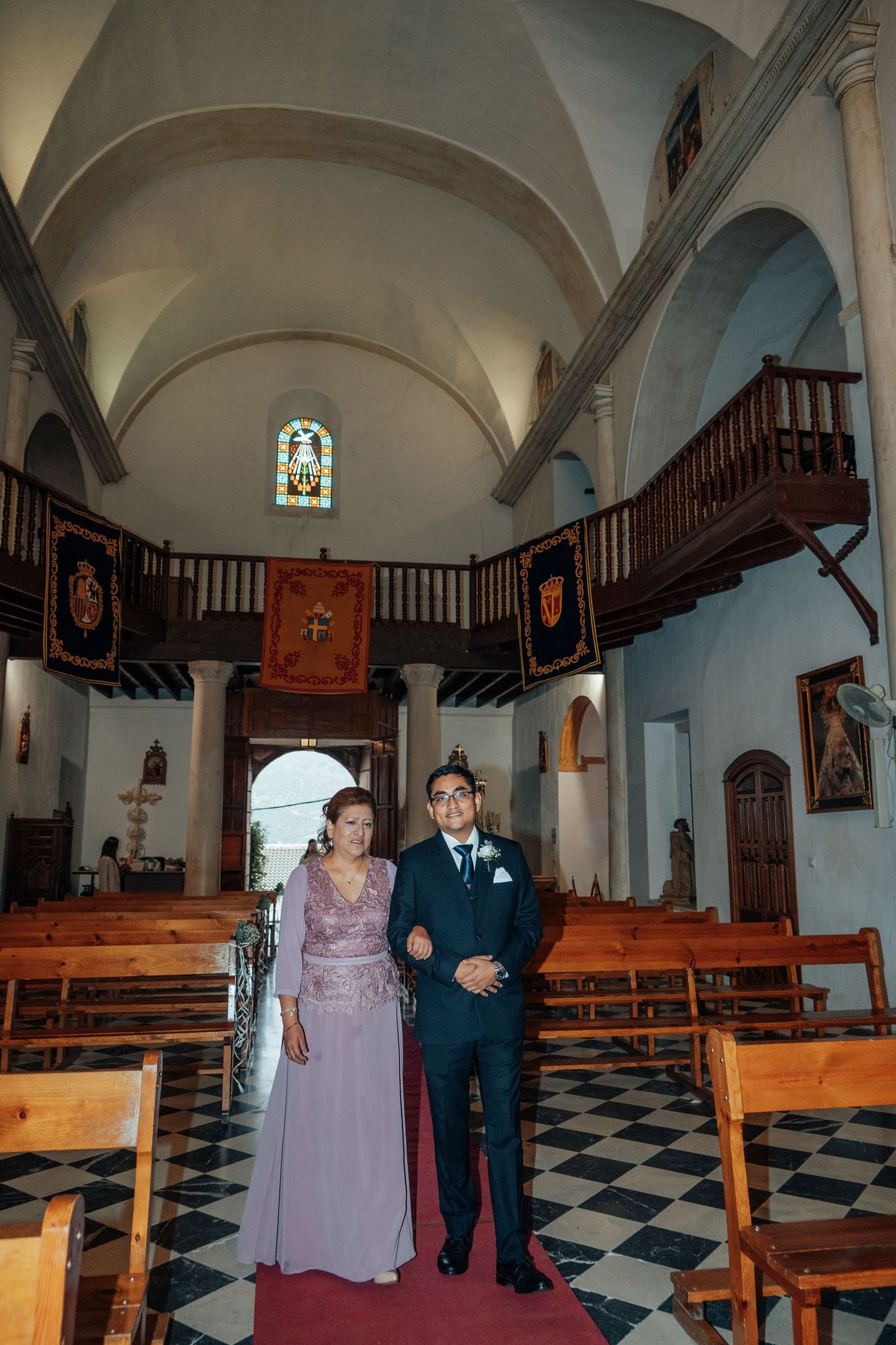 Groom in navy suit walking down church aisle with woman in lavender lace dress during wedding ceremony.
