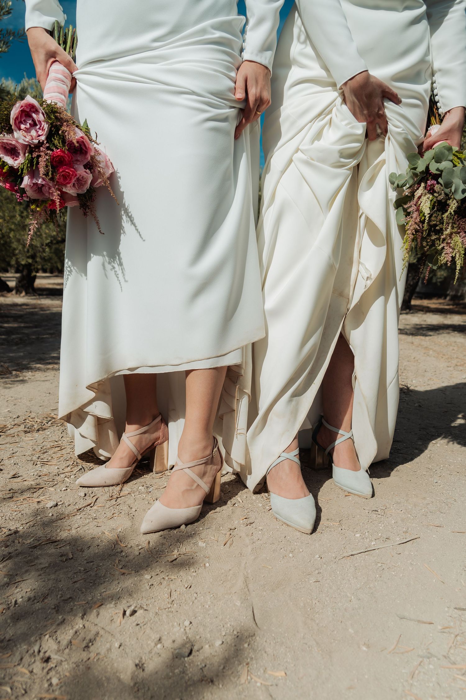 Close-up of two brides in white gowns and heels holding colorful bouquets on sandy ground.
