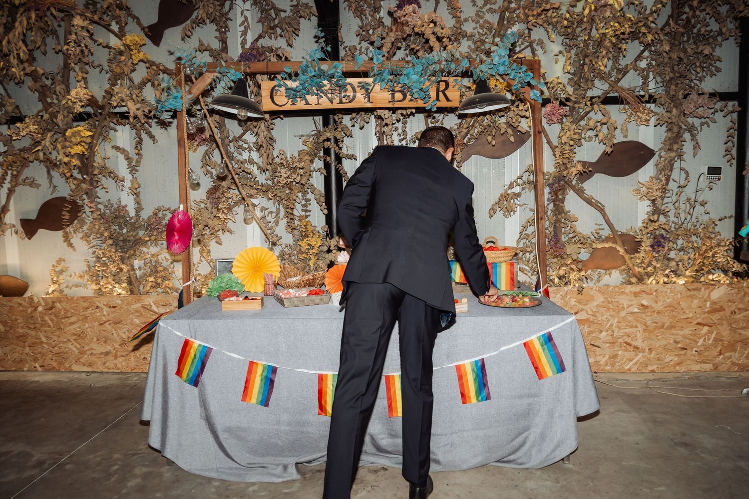 Man in black suit arranging items at a decorated candy bar table with rainbow bunting.