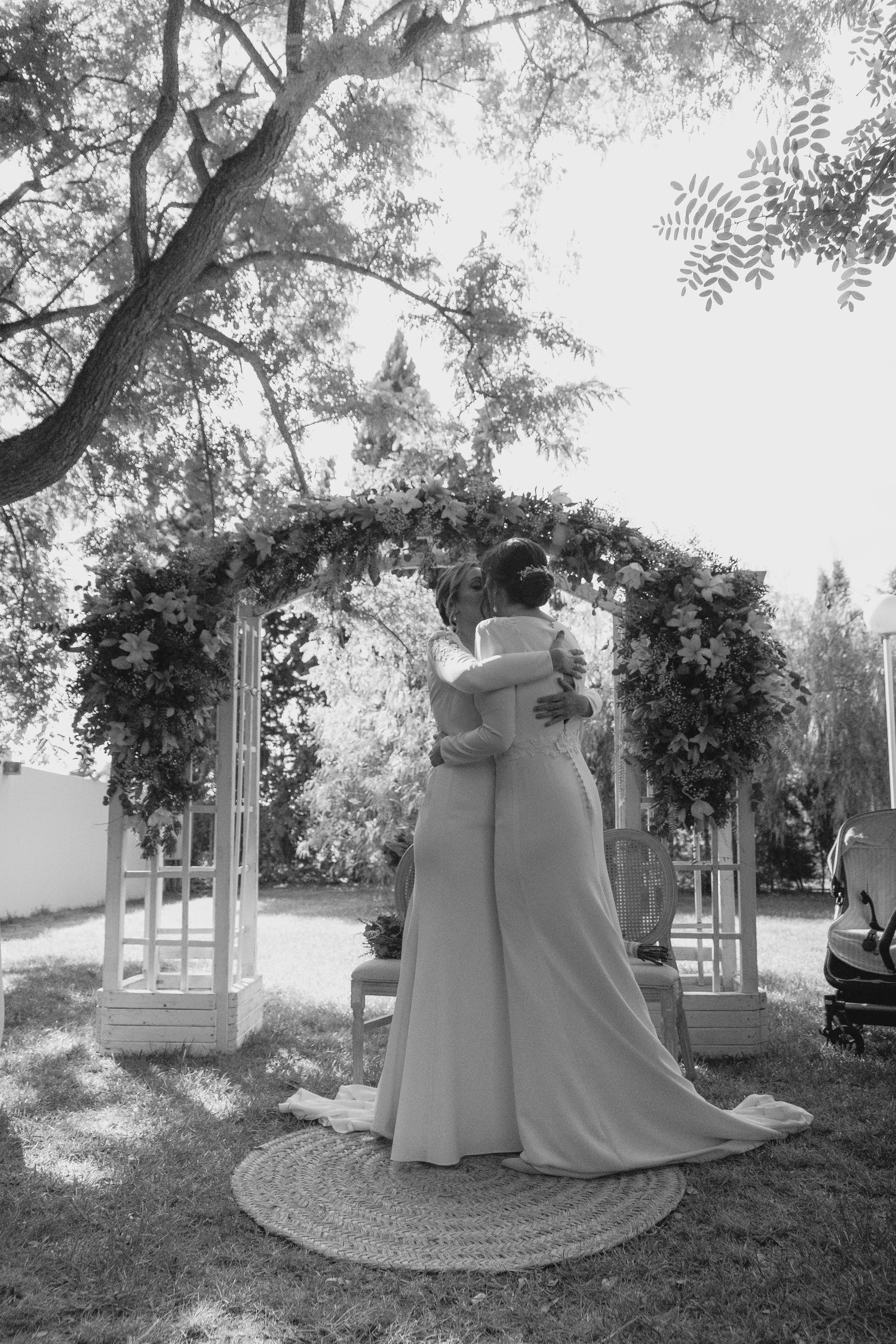 Black and white photo of two brides embracing under floral wedding arch in garden.