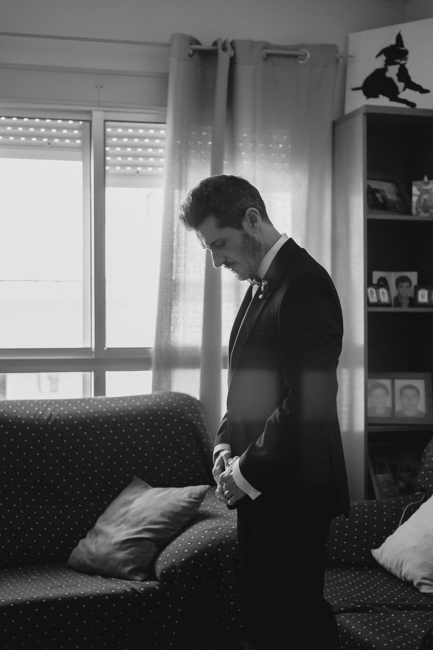 A black and white portrait of a groom in a black suit checking his cufflinks beside a sofa.