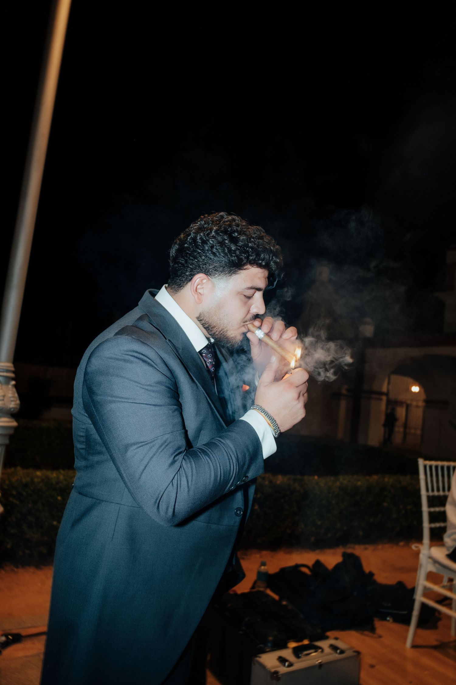 Young man in dark suit lighting a cigar outdoors at night during wedding reception.