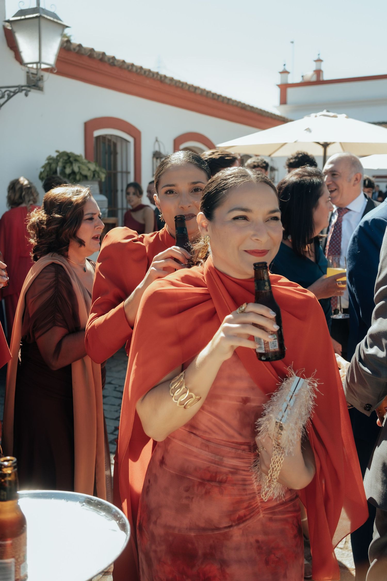 Wedding guest in orange cape dress holds drink while socializing at outdoor Spanish venue reception.