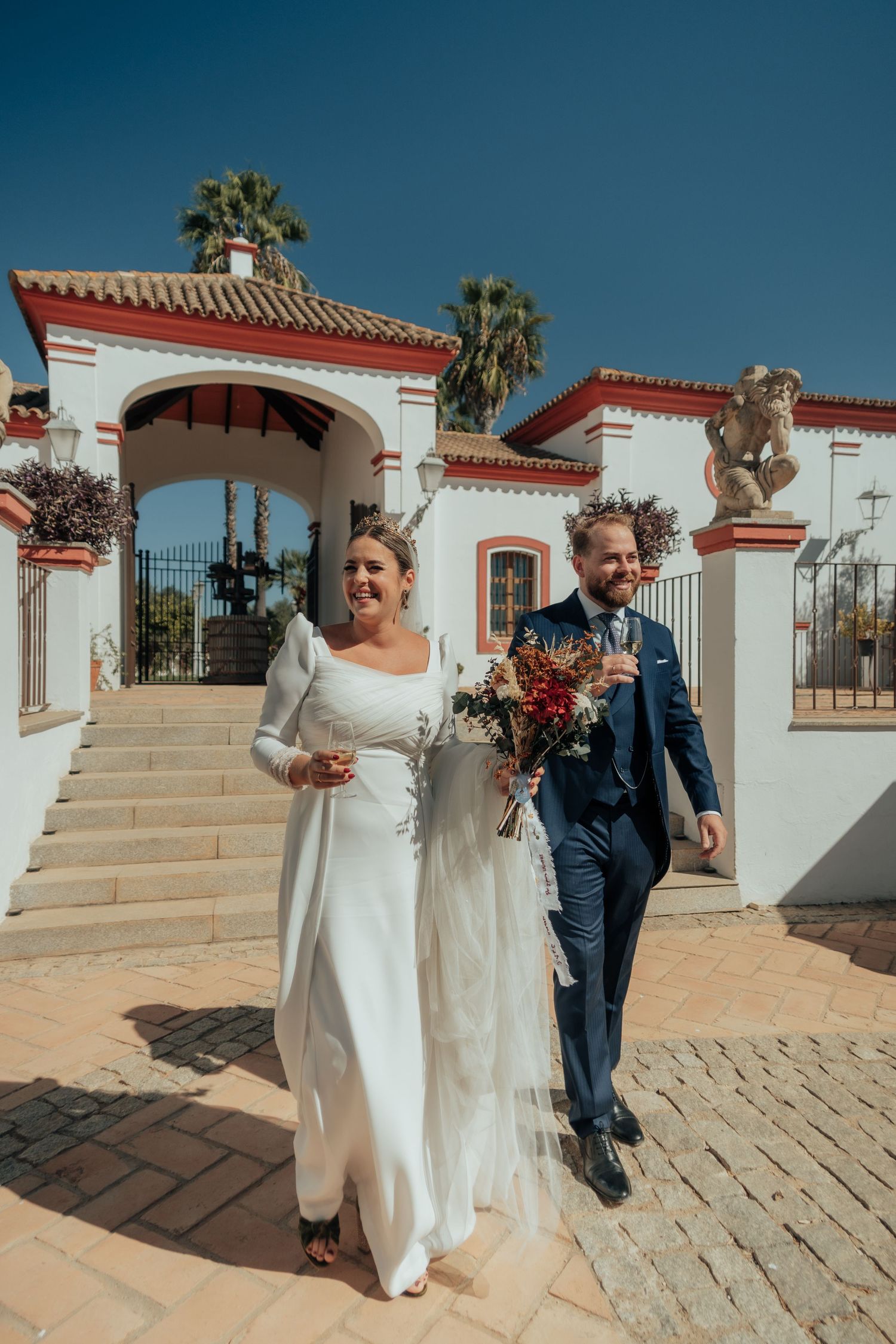 Smiling bride and groom descend steps of elegant white Spanish villa with red trim and palm trees.