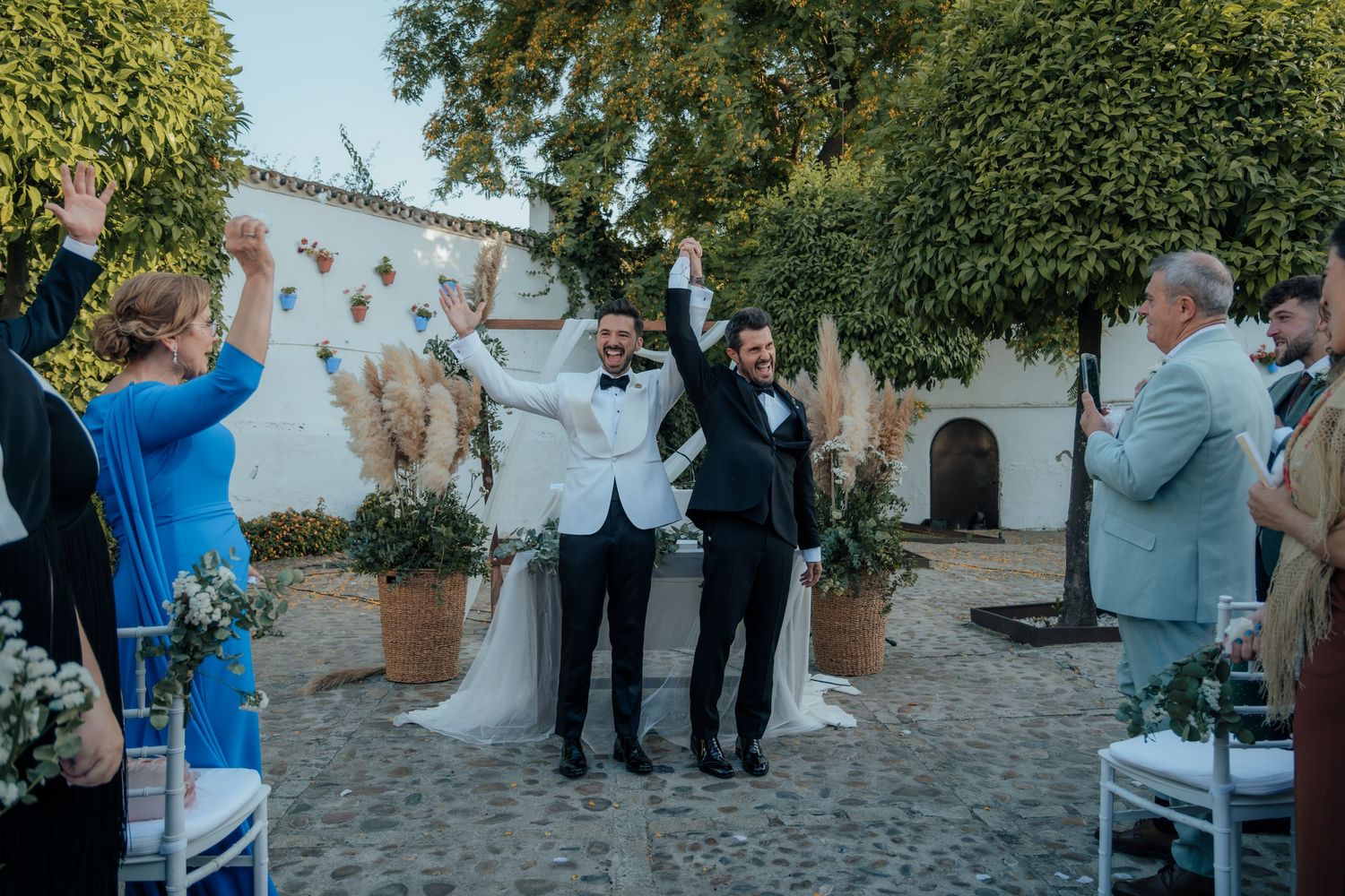 Two grooms celebrate with raised hands walking down aisle as guests cheer at outdoor wedding.
