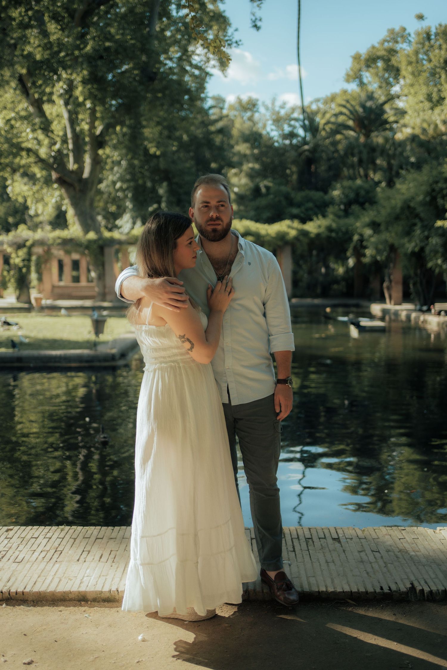 A couple in casual white and grey attire embrace romantically beside a serene green pond in a lush park.