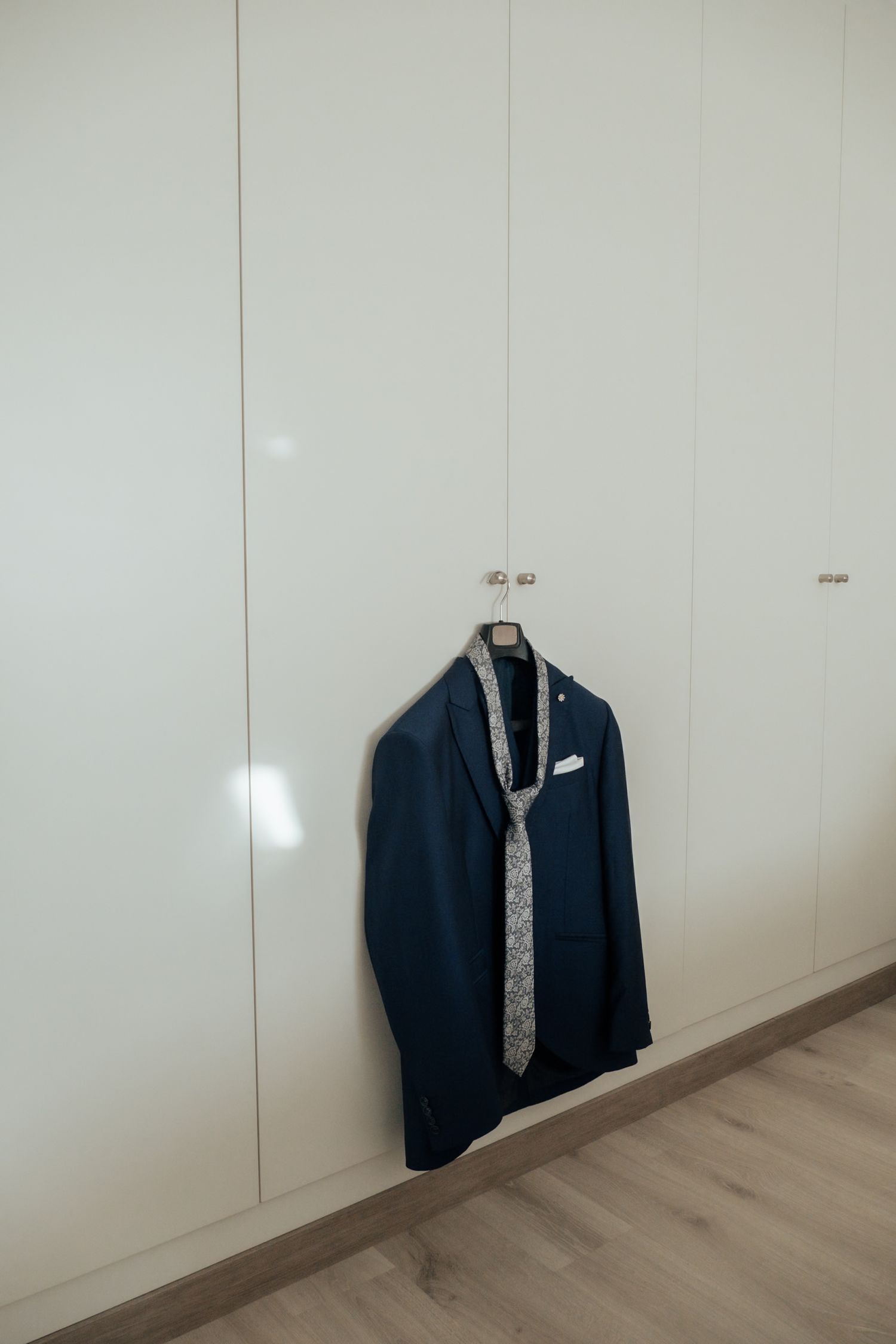 Navy blue wedding suit jacket with a floral tie and white pocket square hanging neatly on a white wardrobe door.