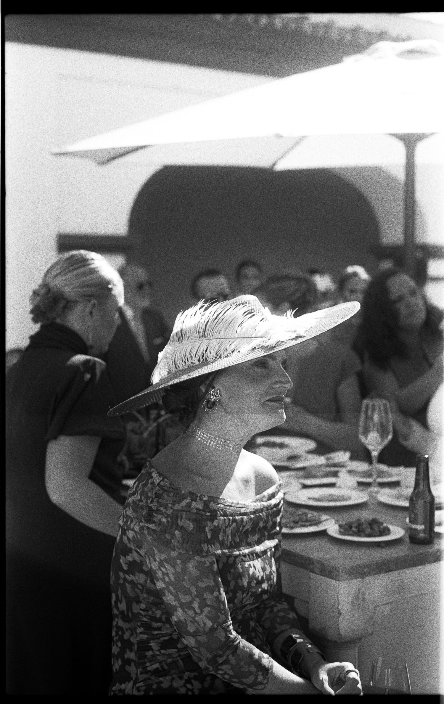 Elegant woman in wide-brim feathered hat laughing at outdoor wedding reception, black and white film photo.
