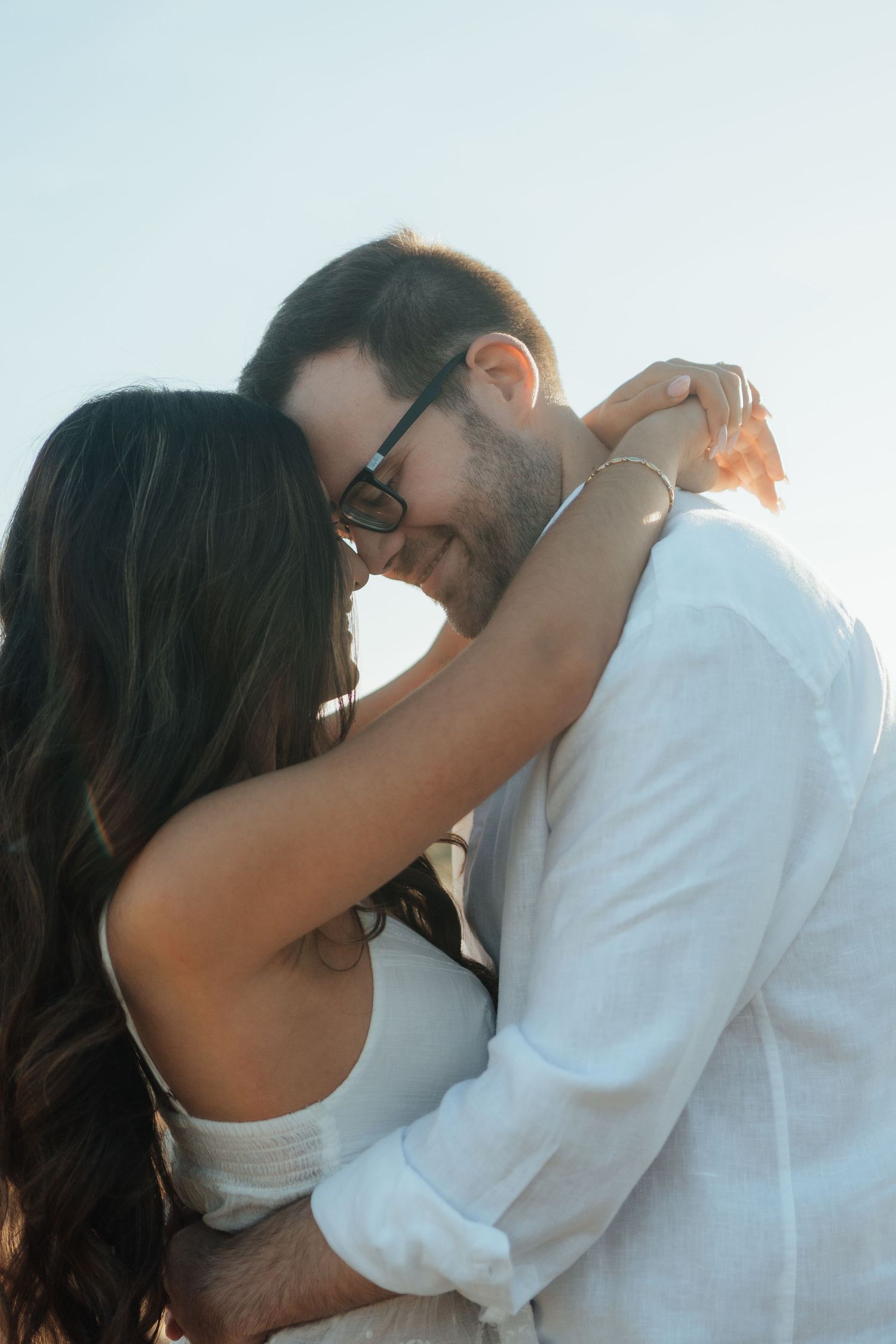 A couple embracing outdoors, man in white shirt and sunglasses smiling as woman with long dark hair wraps arms around his neck.
