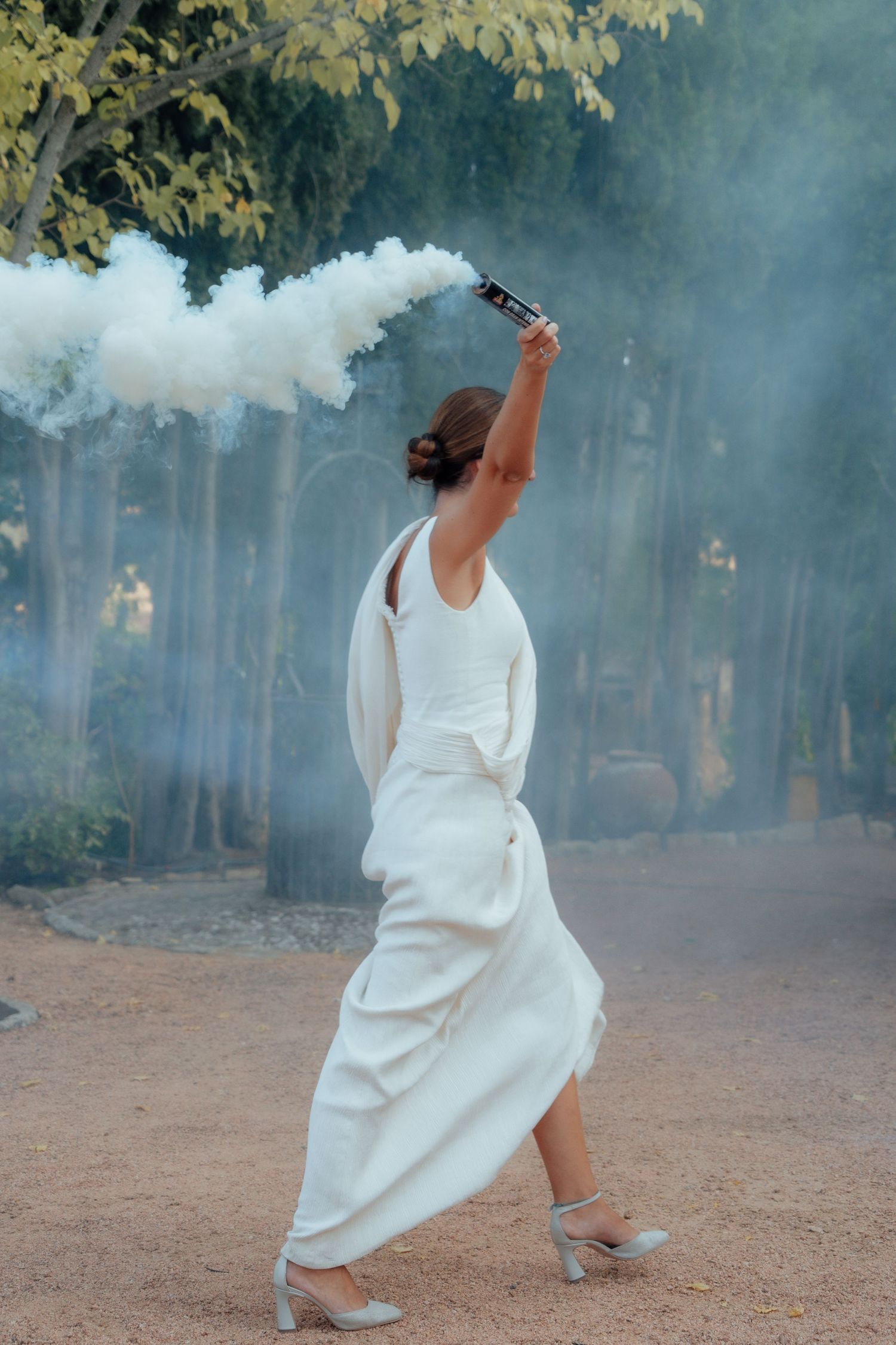 Bride in white gown holding white smoke flare walking through tree-lined path at wedding.