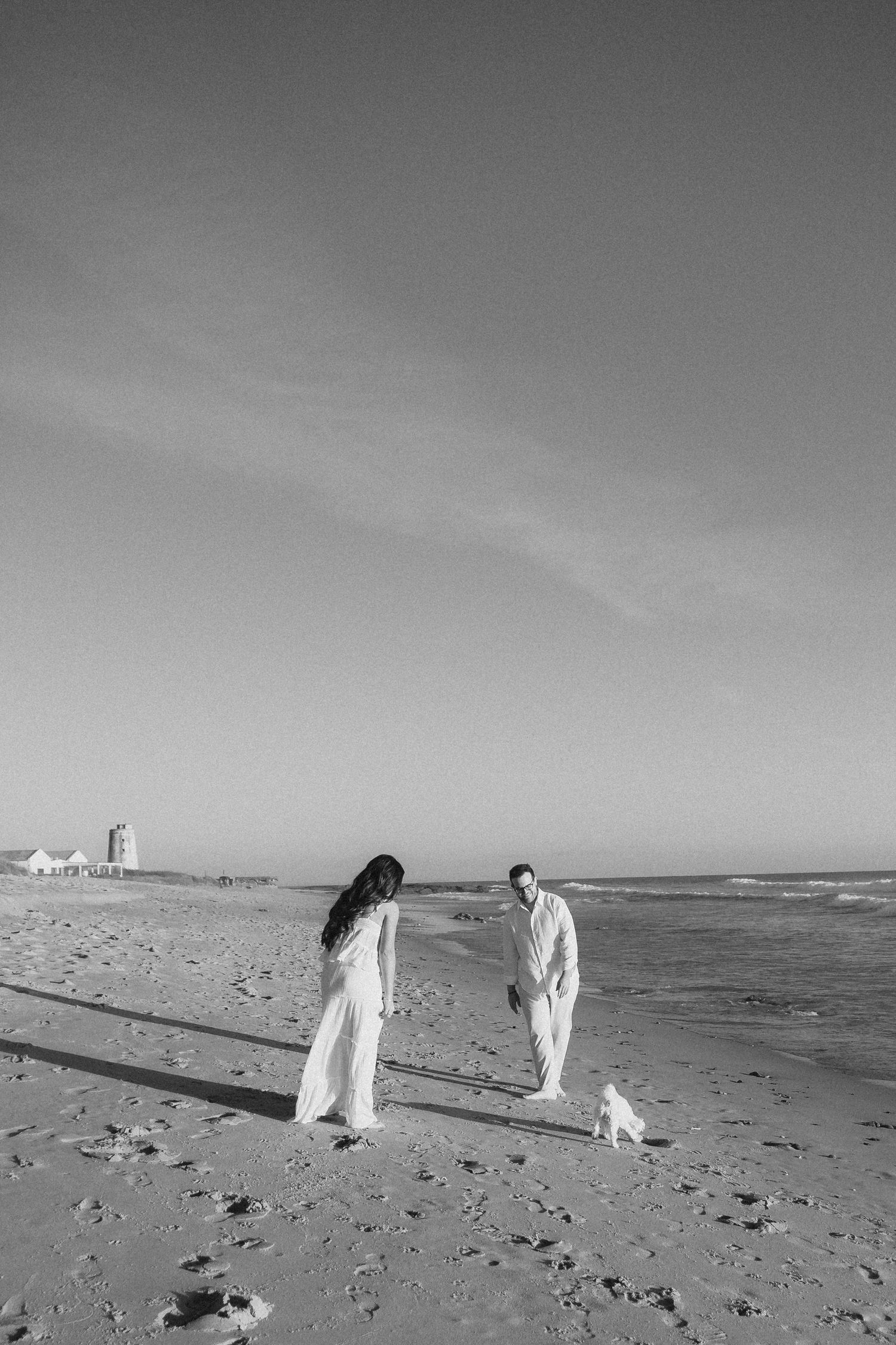 Black and white photo of a couple in white attire playing with a small dog on a sandy beach with ocean waves.