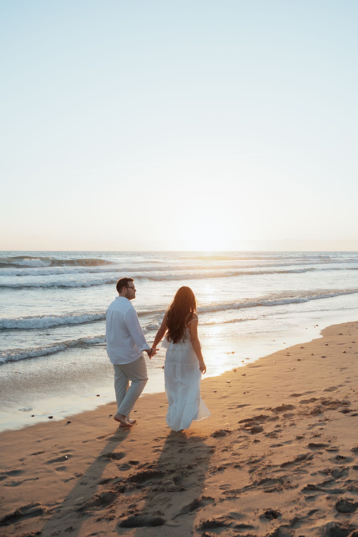 Couple in white outfits holding hands walking along a sandy beach at sunset with ocean waves in the background.