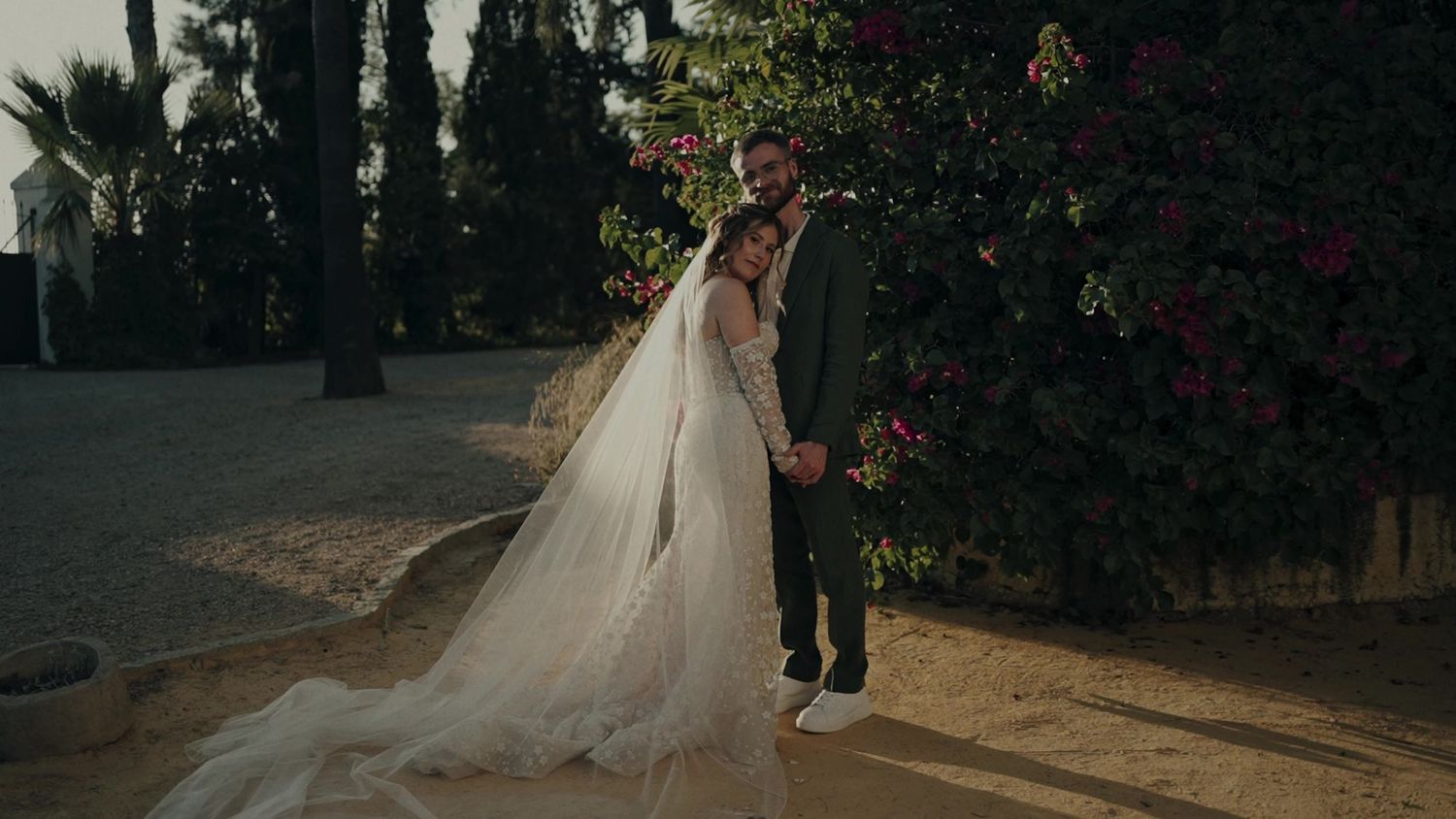 Bride in flowing tulle gown embraces groom beside vibrant bougainvillea at sunset wedding.