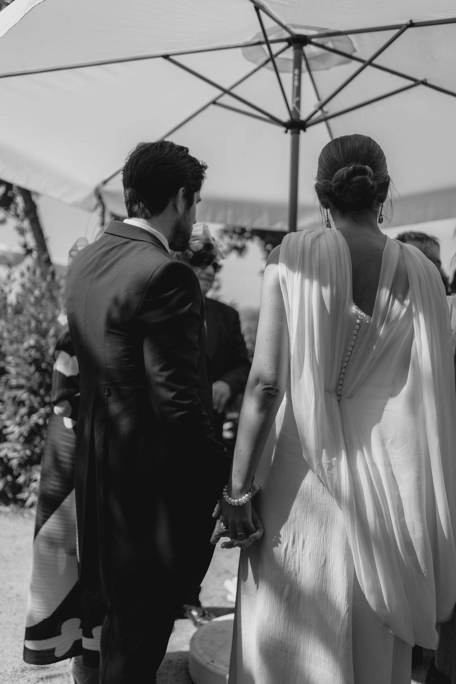Black and white photo of bride and groom walking hand in hand under white parasol at wedding.