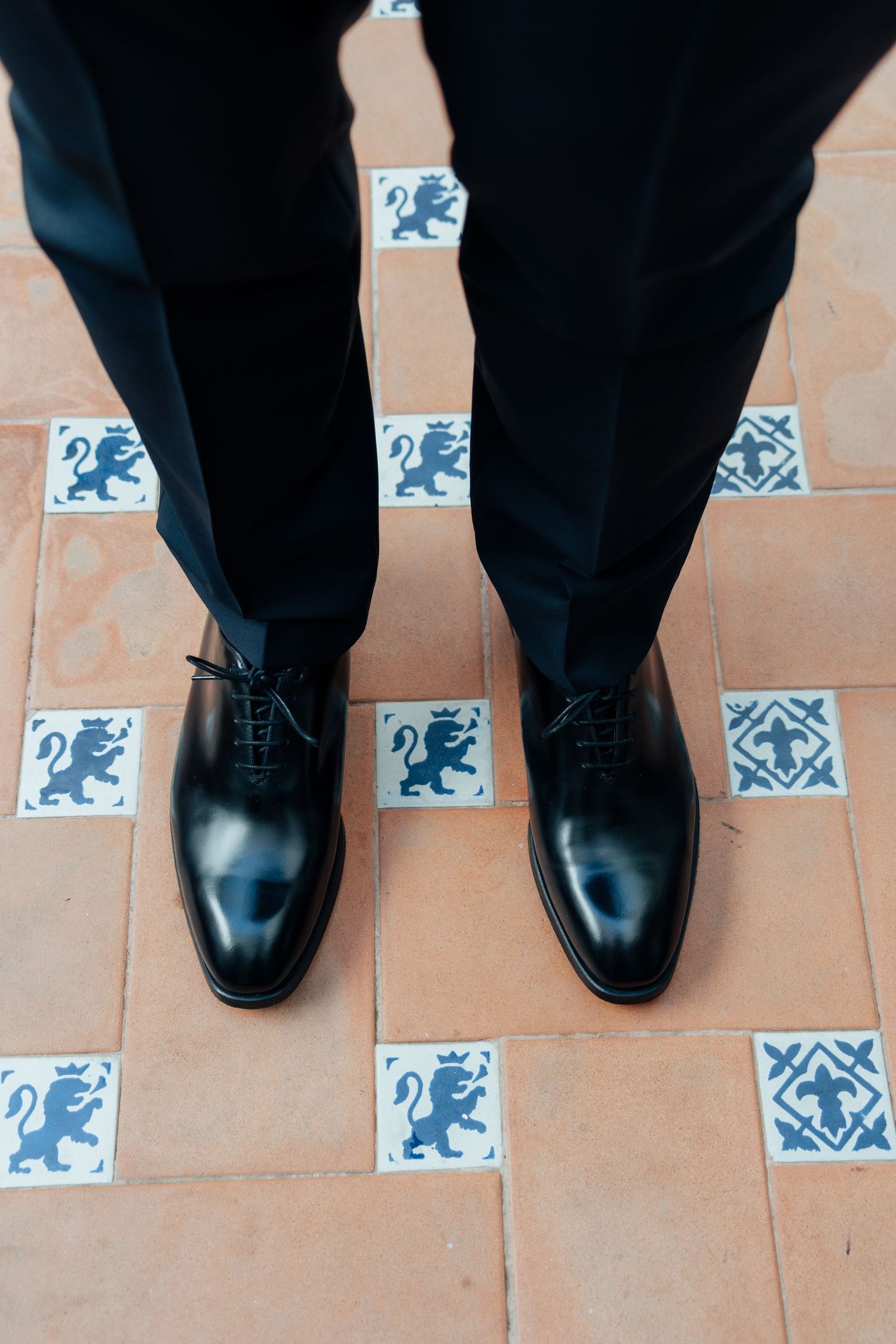 Top view of groom's polished black Oxford shoes on decorative terracotta tiles with blue lion motifs.