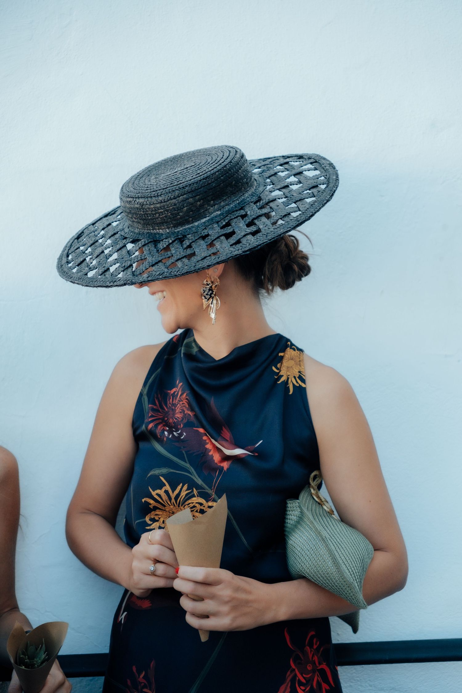 Elegant wedding guest wearing a wide-brim woven hat and floral dress holding a confetti cone outdoors.