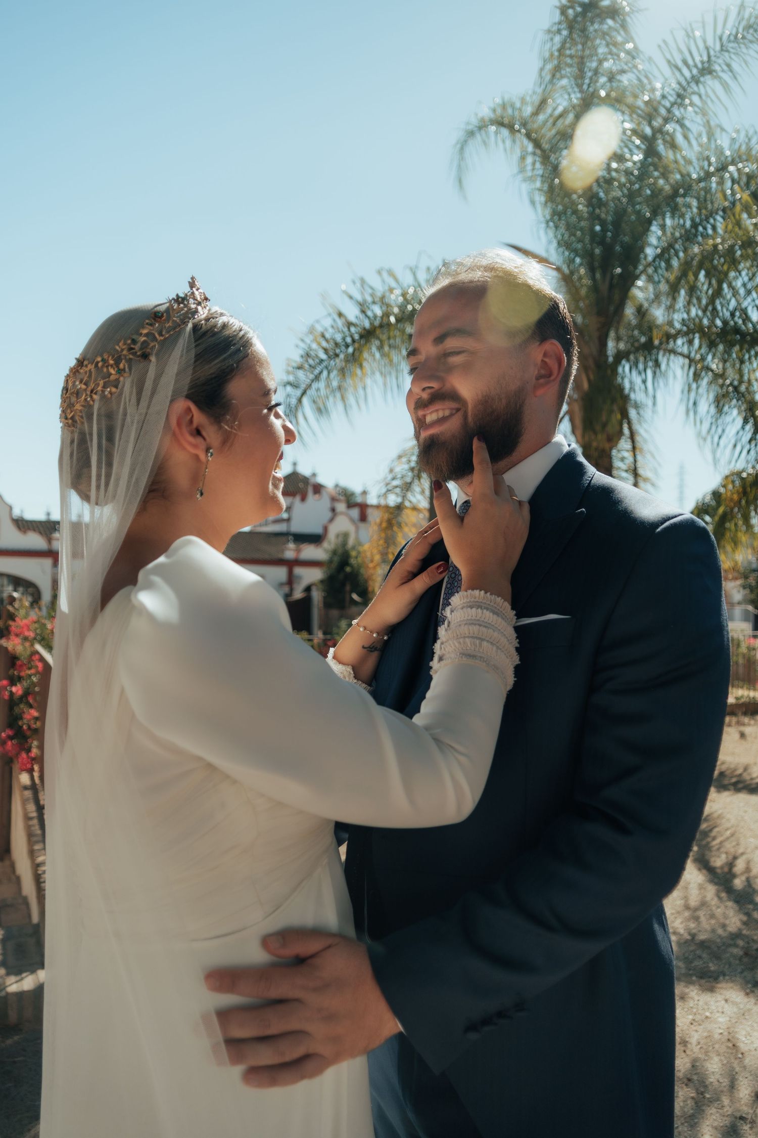 Bride in white gown tenderly touches bearded groom's face as they smile at each other outdoors.