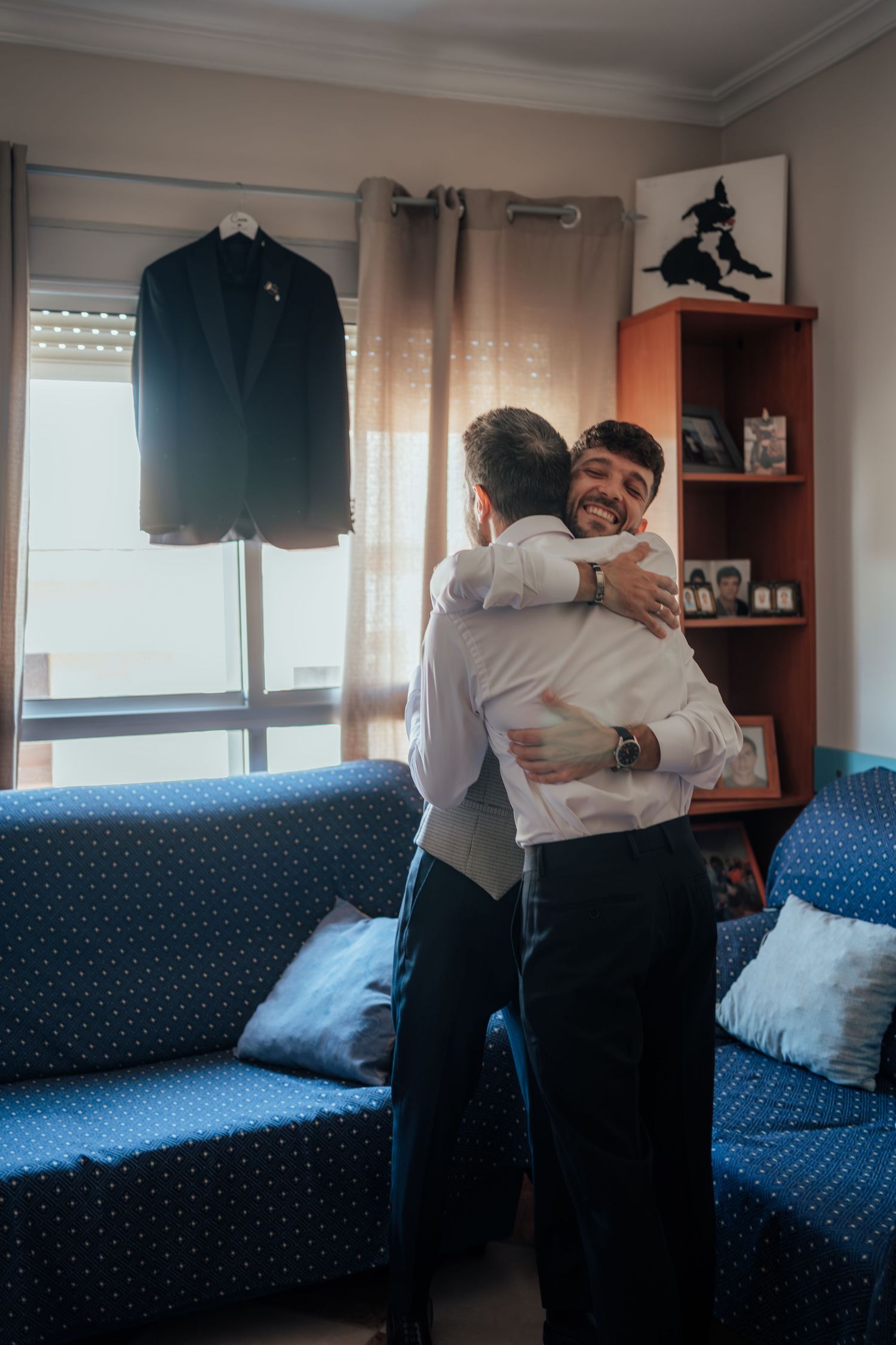 Two men share a warm hug in a living room with a black suit jacket hanging in the background.