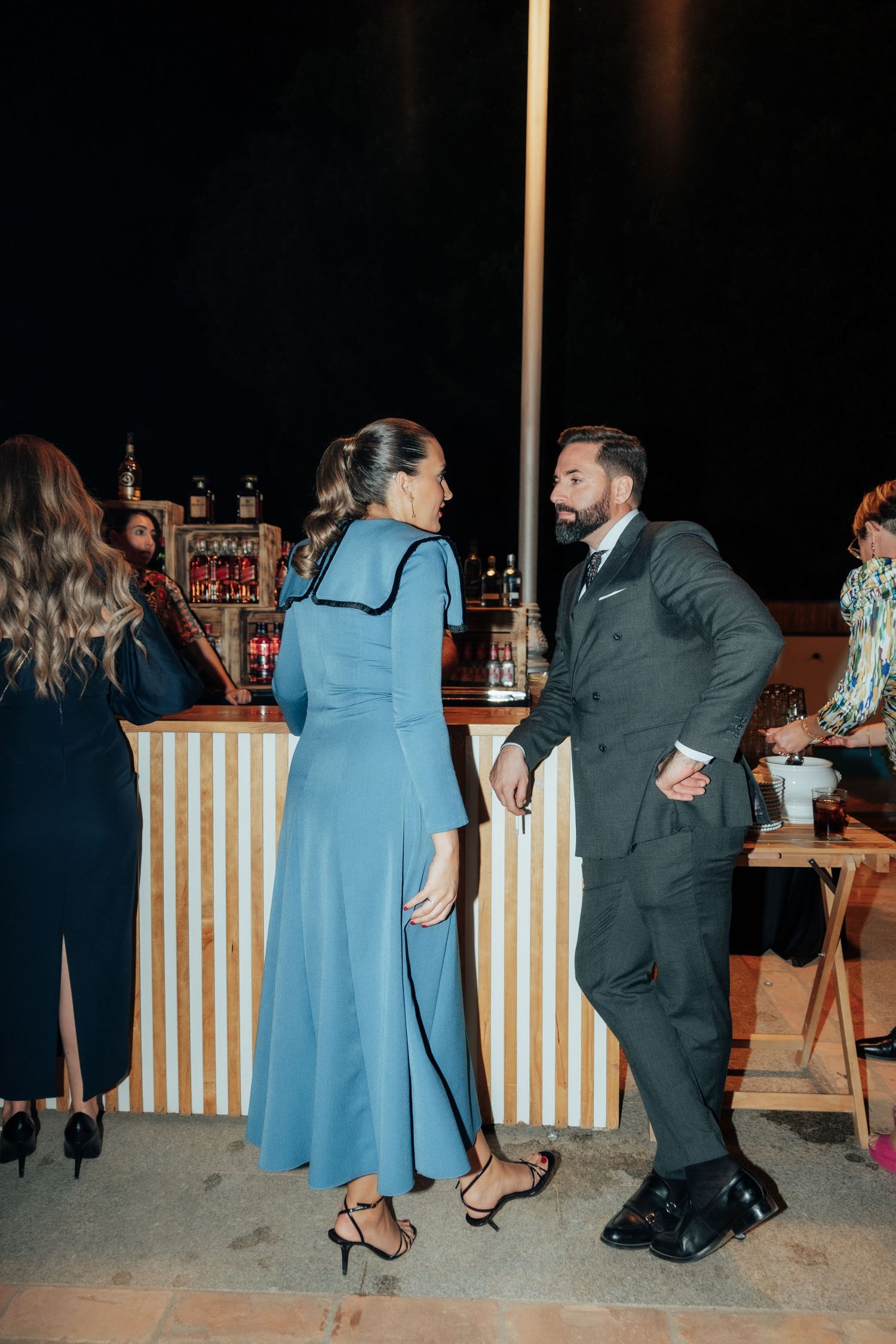Man and woman in elegant attire conversing near a striped outdoor bar at night.