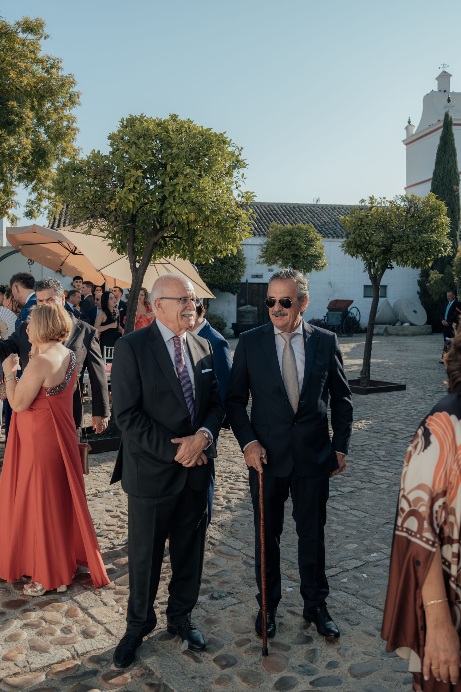 Two elderly gentlemen in suits chat together at an outdoor wedding venue on a sunny day.