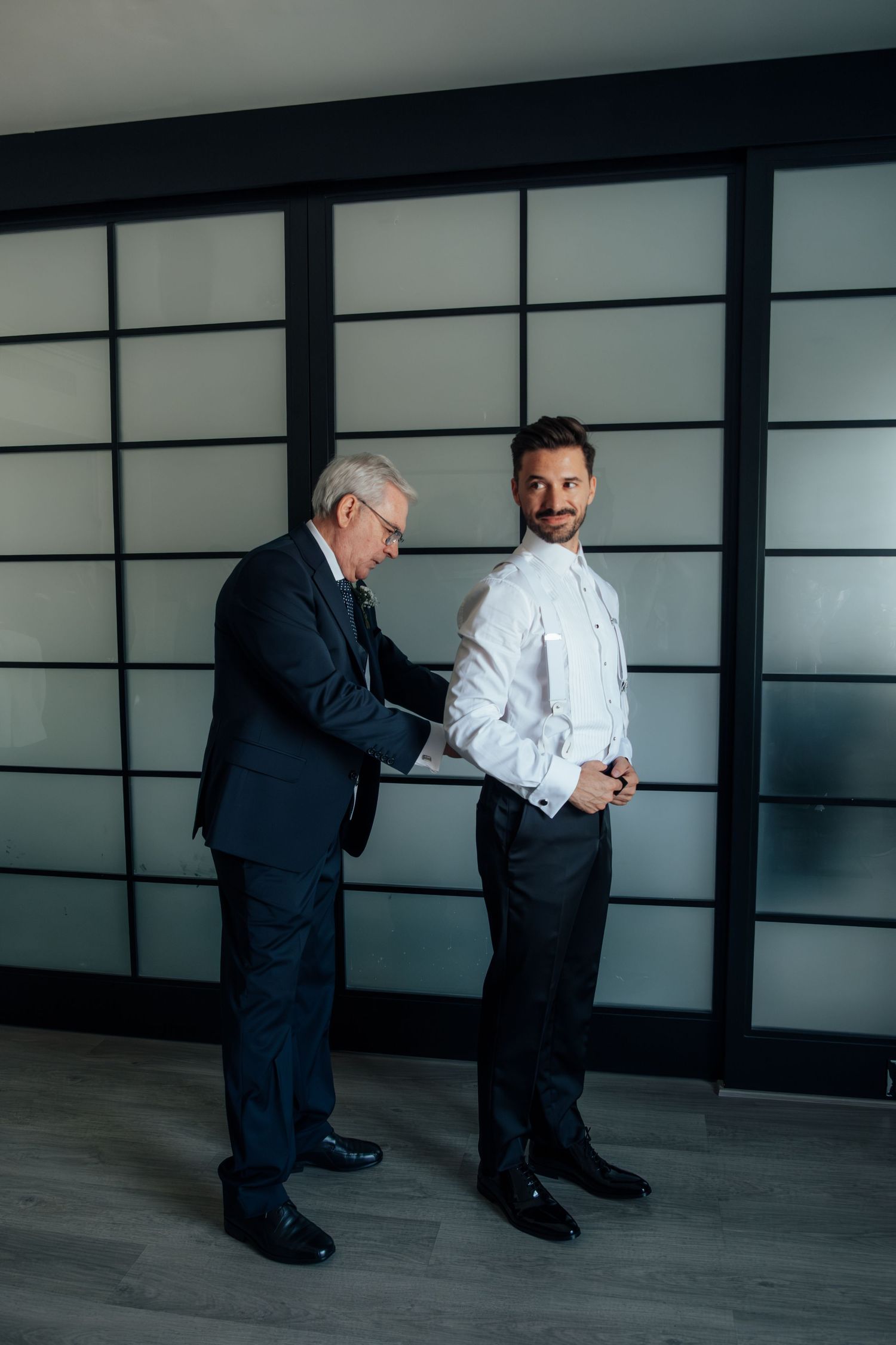 An older man in a navy suit helps a younger man button his white tuxedo shirt indoors.